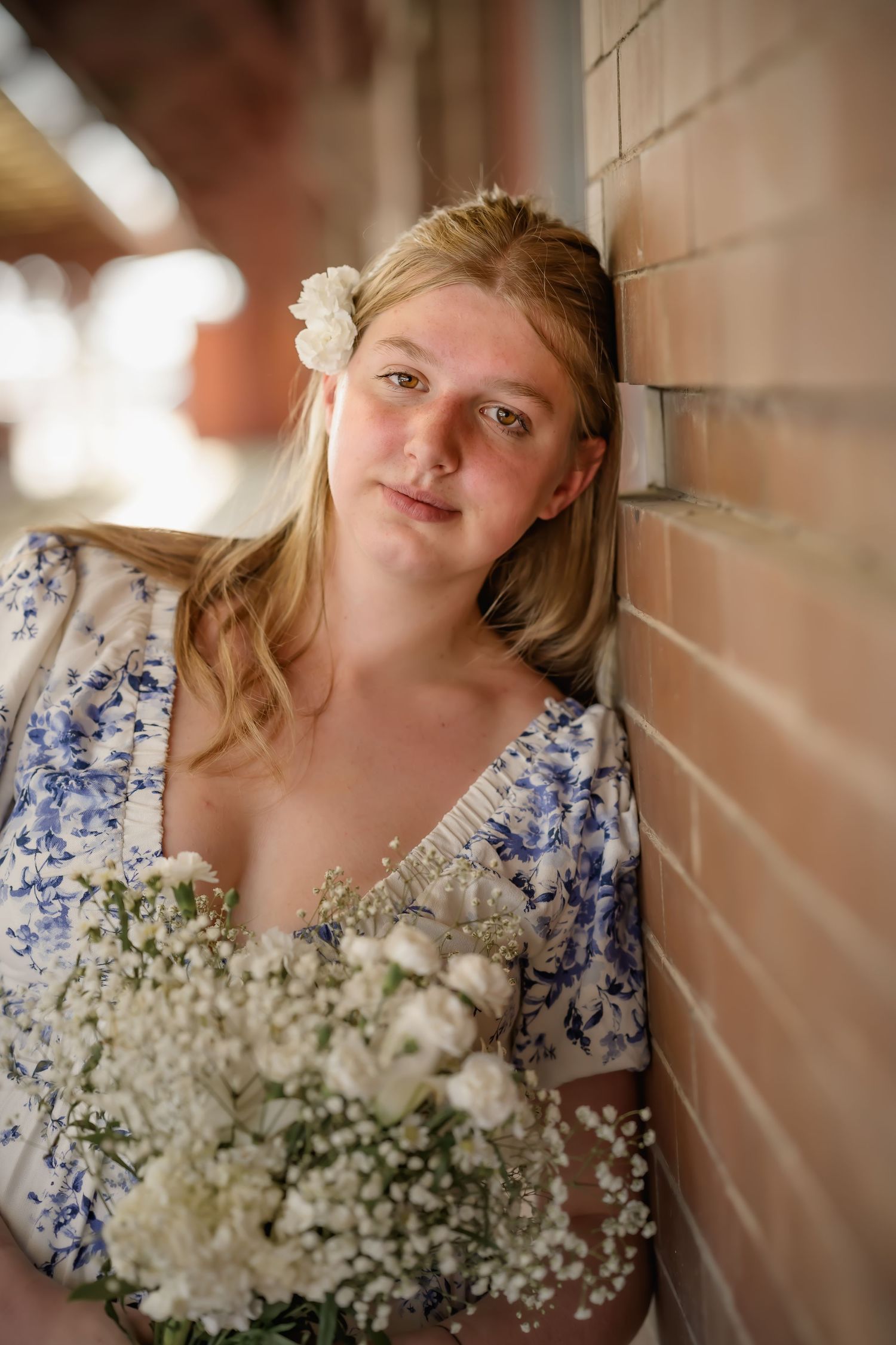 Blonde teen girl in blue floral dress holds white flower bouquet while leaning against a brick wall.