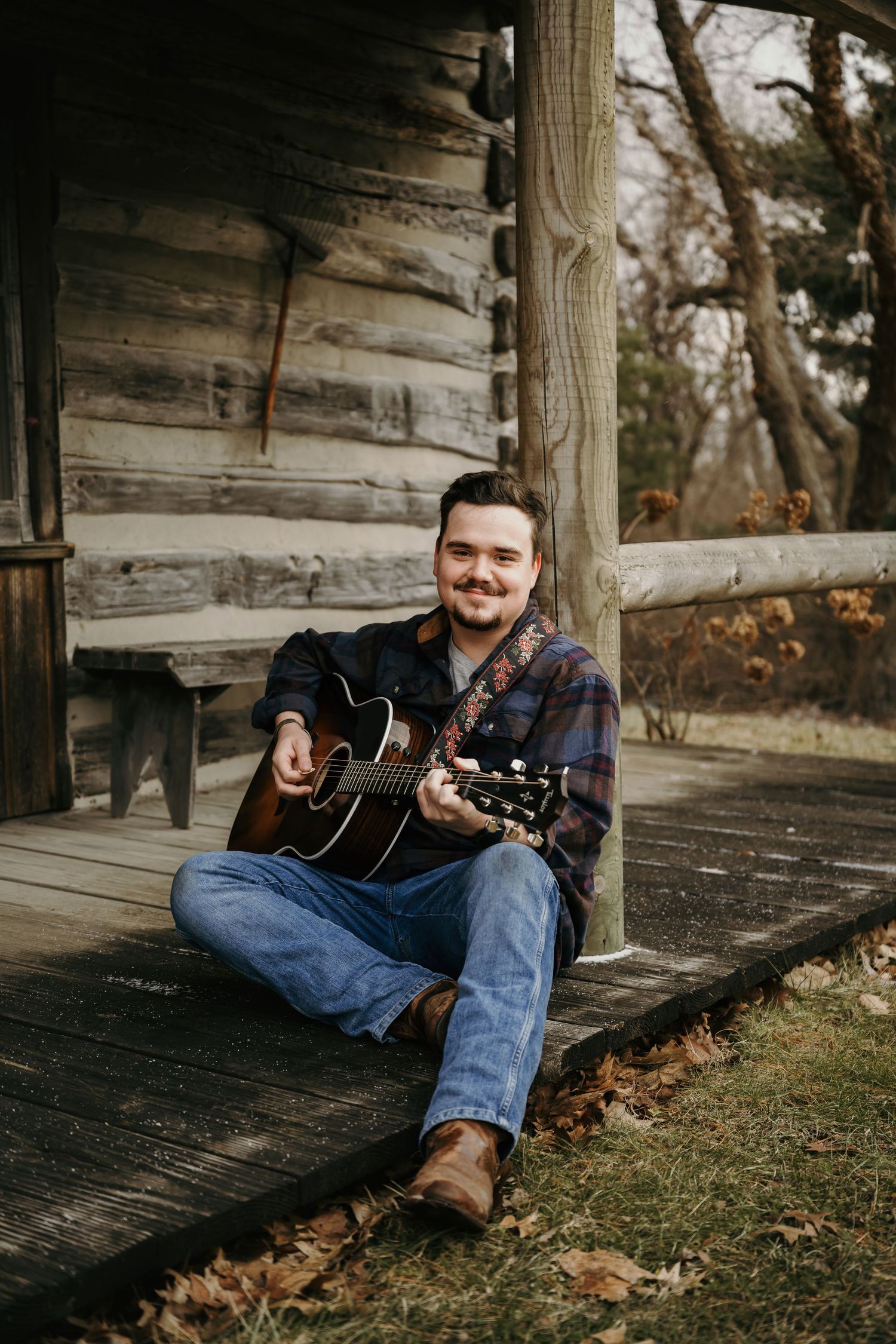 Young man playing acoustic guitar while sitting on the porch steps of a rustic log cabin.