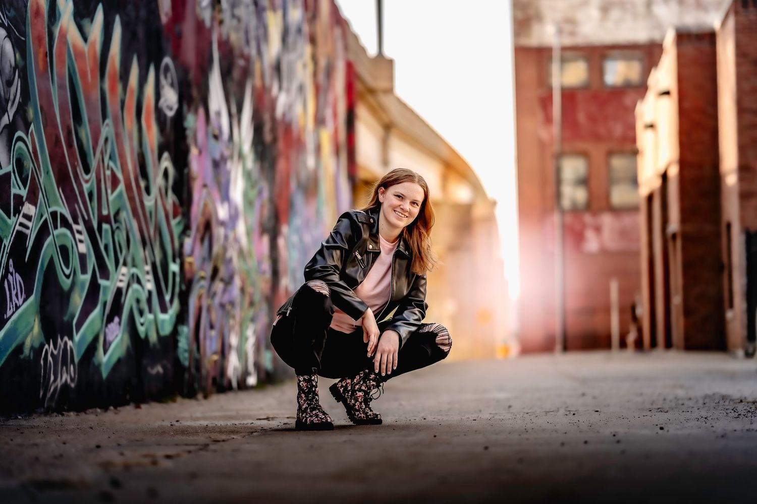 Smiling young woman squatting in urban alley beside vibrant graffiti wall with golden sunflare.