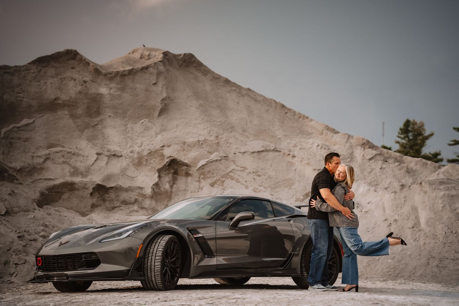 Romantic couple embraces beside a gray Corvette sports car against a sandy quarry backdrop.