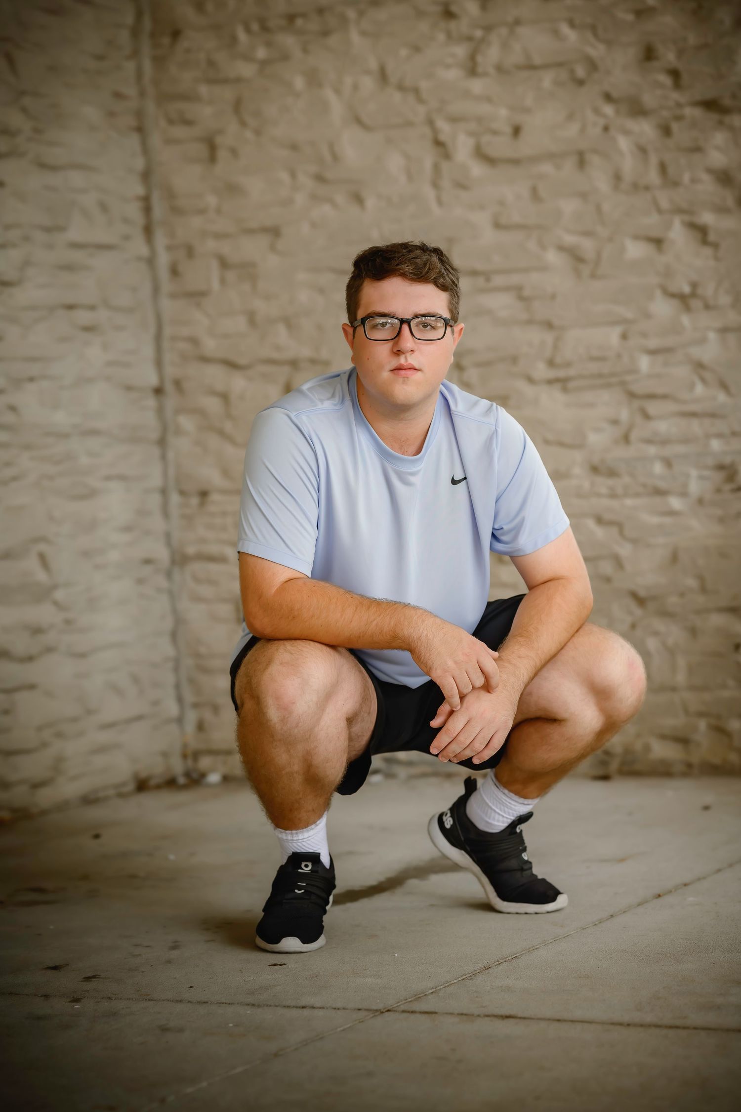 Teen boy in light blue Nike shirt crouches in front of stone wall wearing glasses.