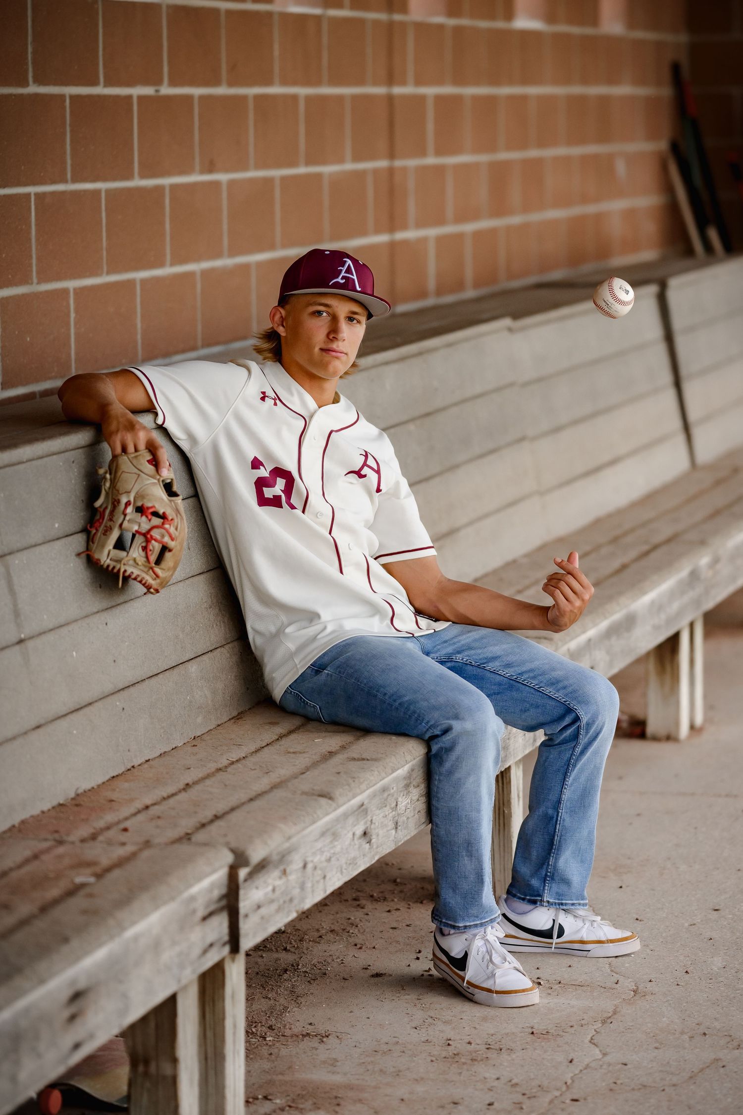 High school baseball player in maroon uniform sits in dugout tossing a ball while wearing a team cap and Nike shoes.