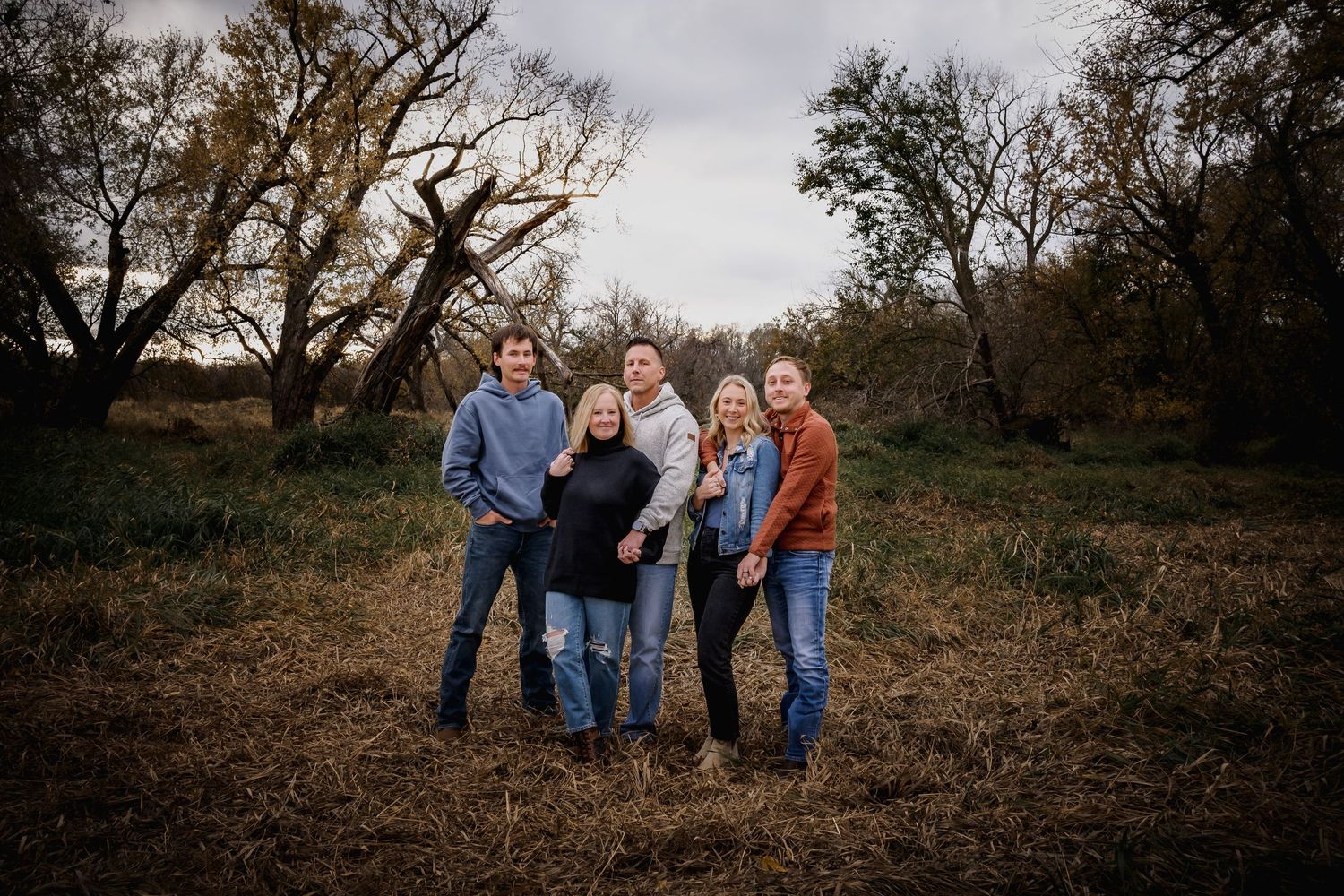 Family of five posing together in a rustic outdoor field with bare trees in fall.