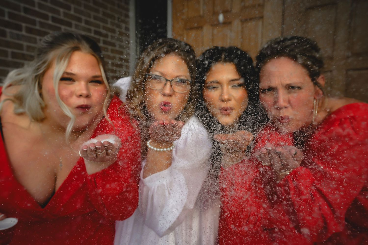Four women in red dresses and one in white blow glitter toward camera in fun celebration photo.