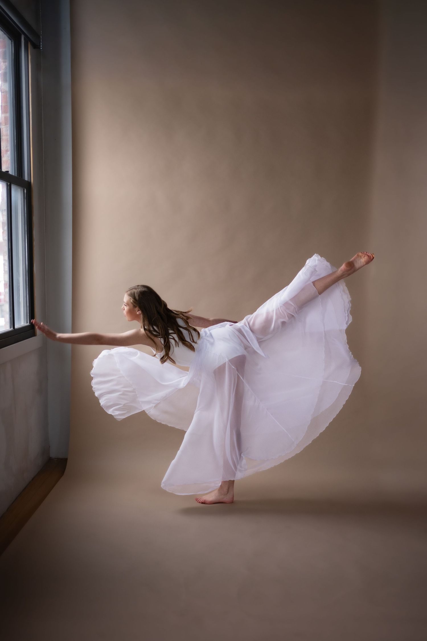 Graceful dancer in flowing white dress performing an arabesque pose near a studio window.
