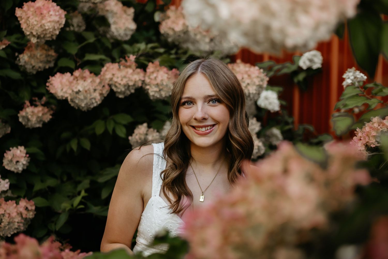 Smiling teenage girl poses among pink and white hydrangea blooms in a garden setting.
