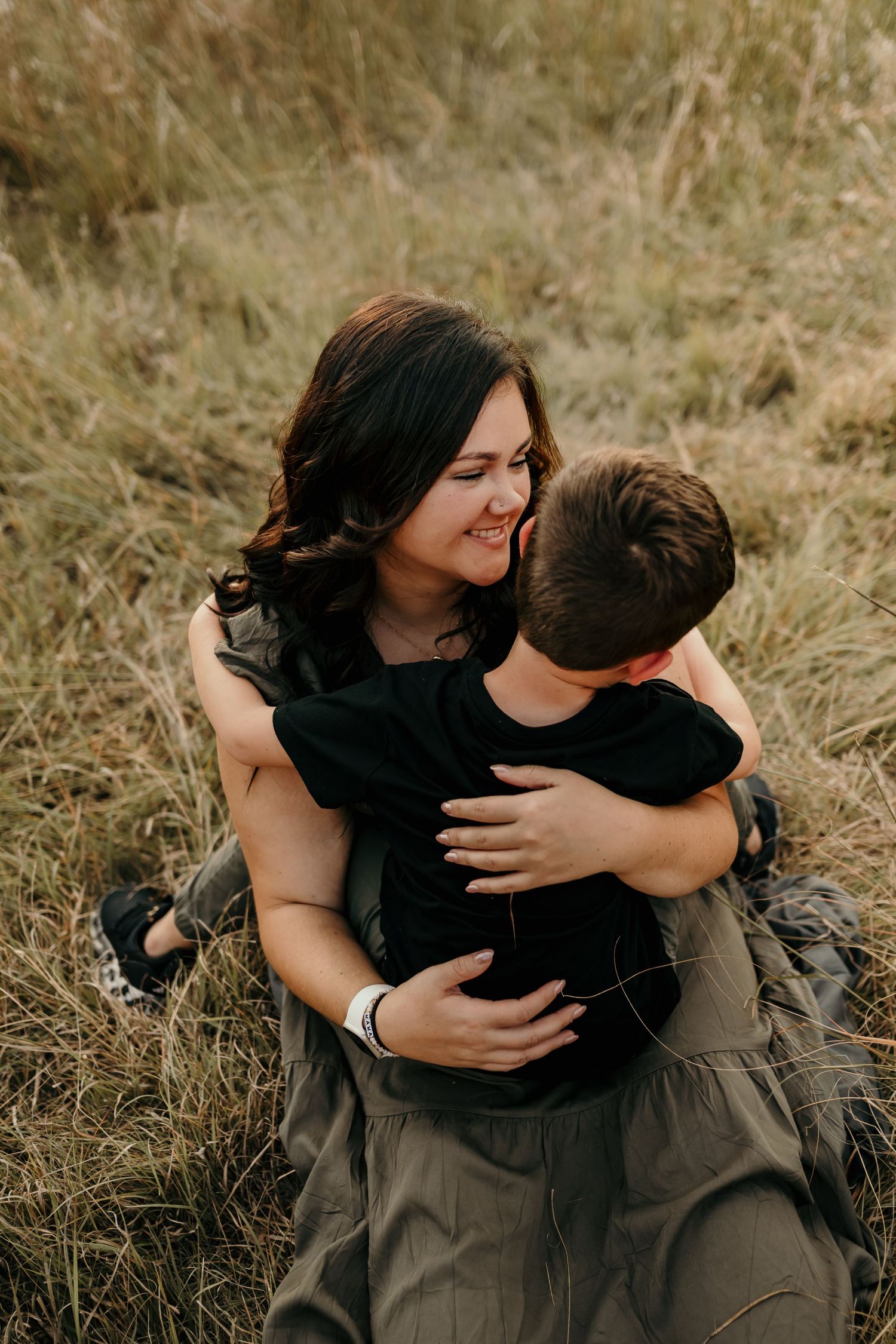 A woman smiles joyfully while hugging a young boy in a grassy field, sharing a warm and loving outdoor moment.