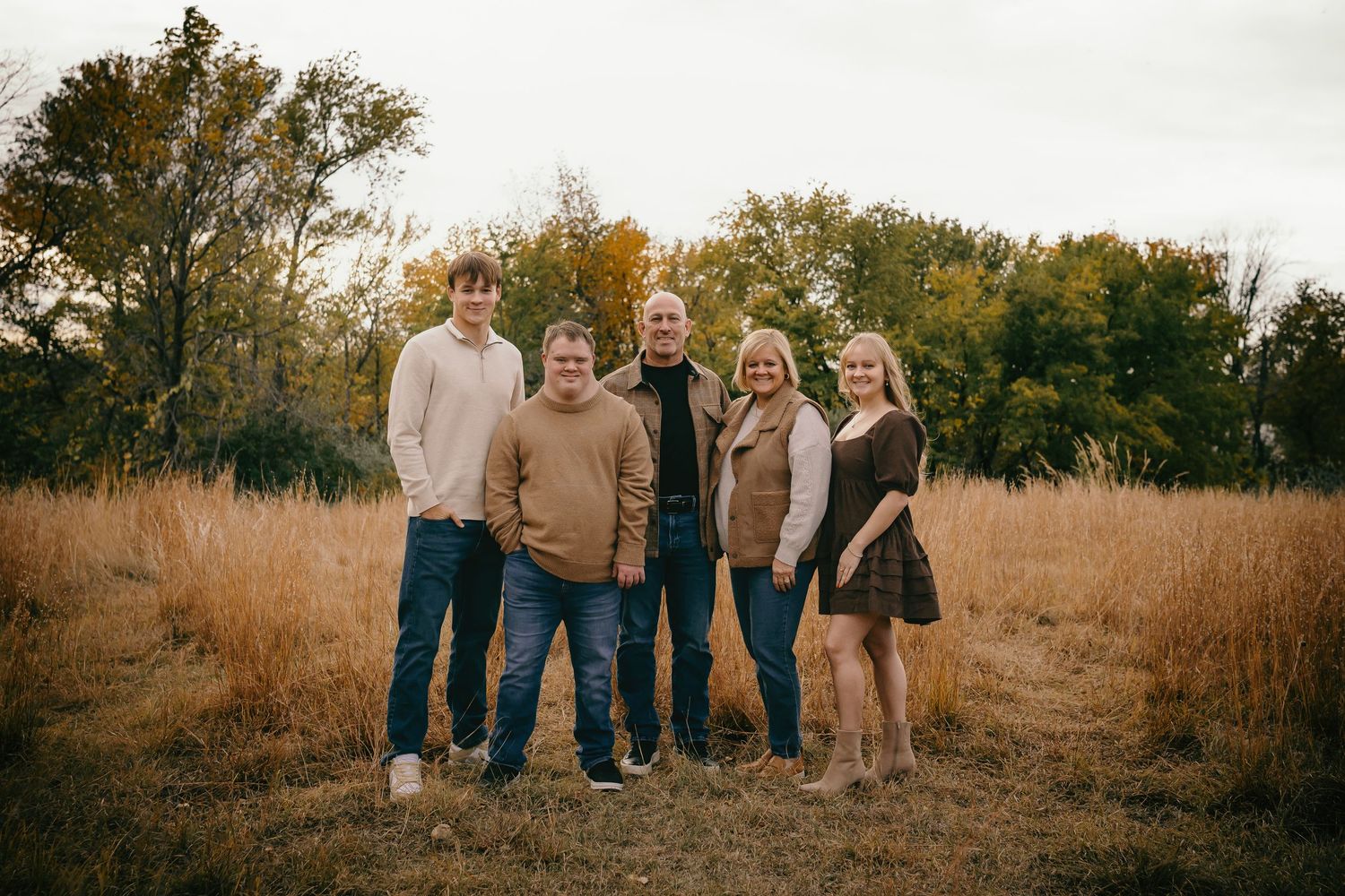 Family of five posing in a golden autumn field with trees in the background.