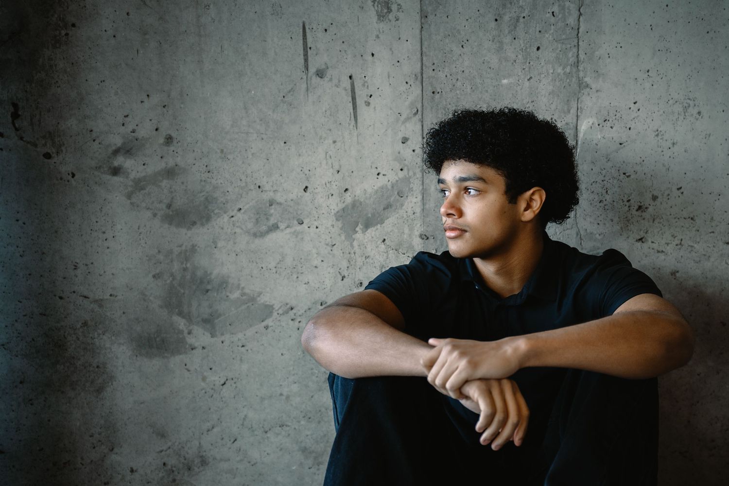 Thoughtful young man with curly hair sitting against a concrete wall with arms crossed on knees.