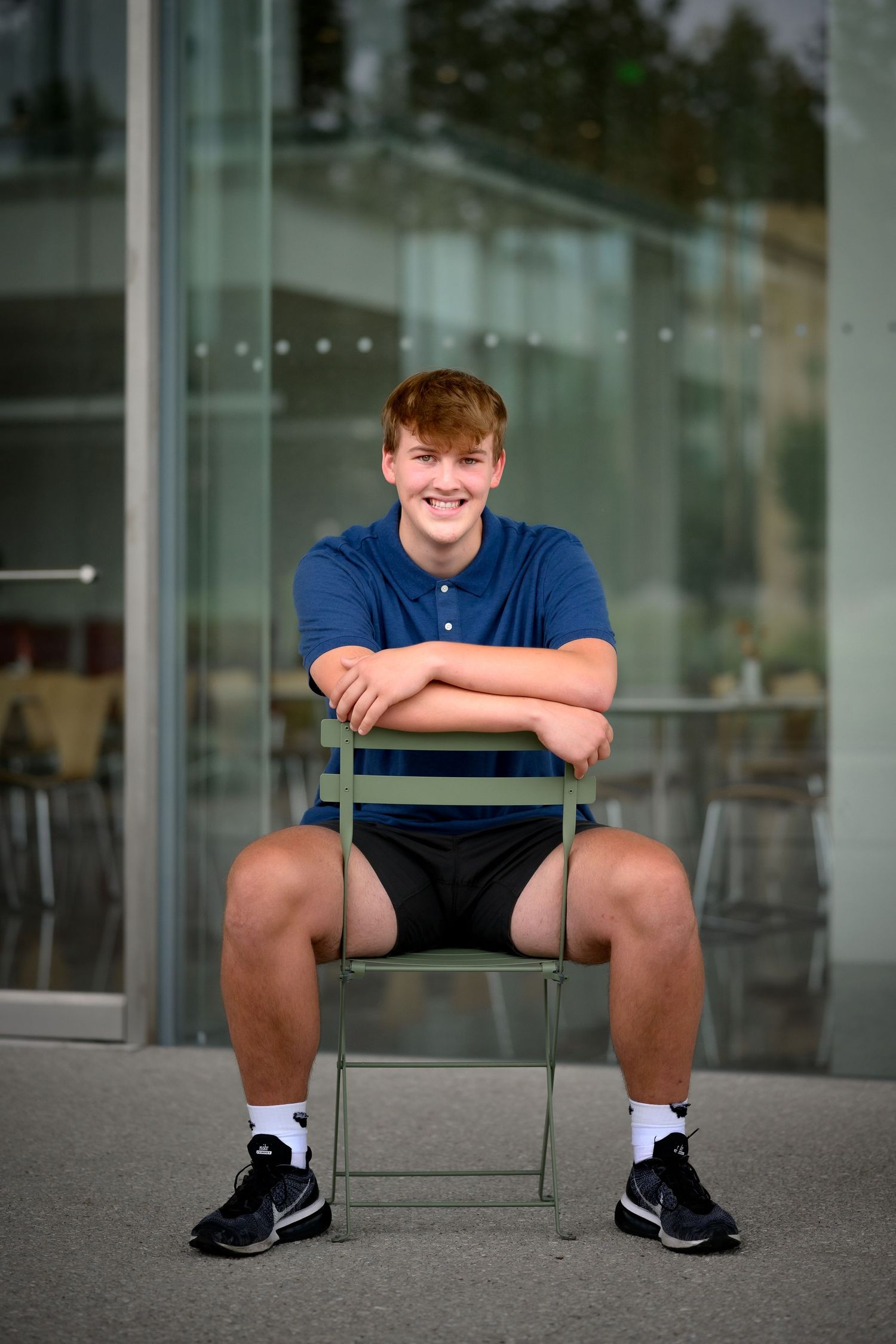 Smiling redhead teen boy in blue polo sitting on green chair outside glass building.