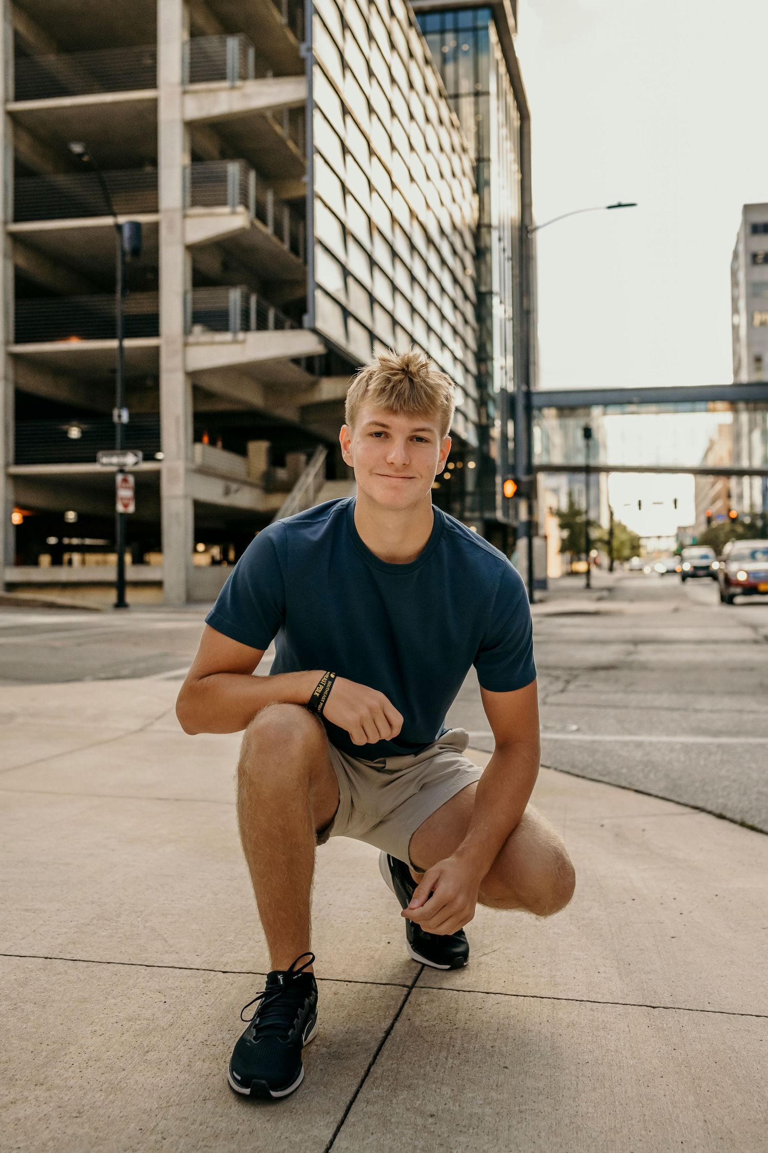 Teen boy in navy shirt crouches on urban sidewalk with city buildings in background.