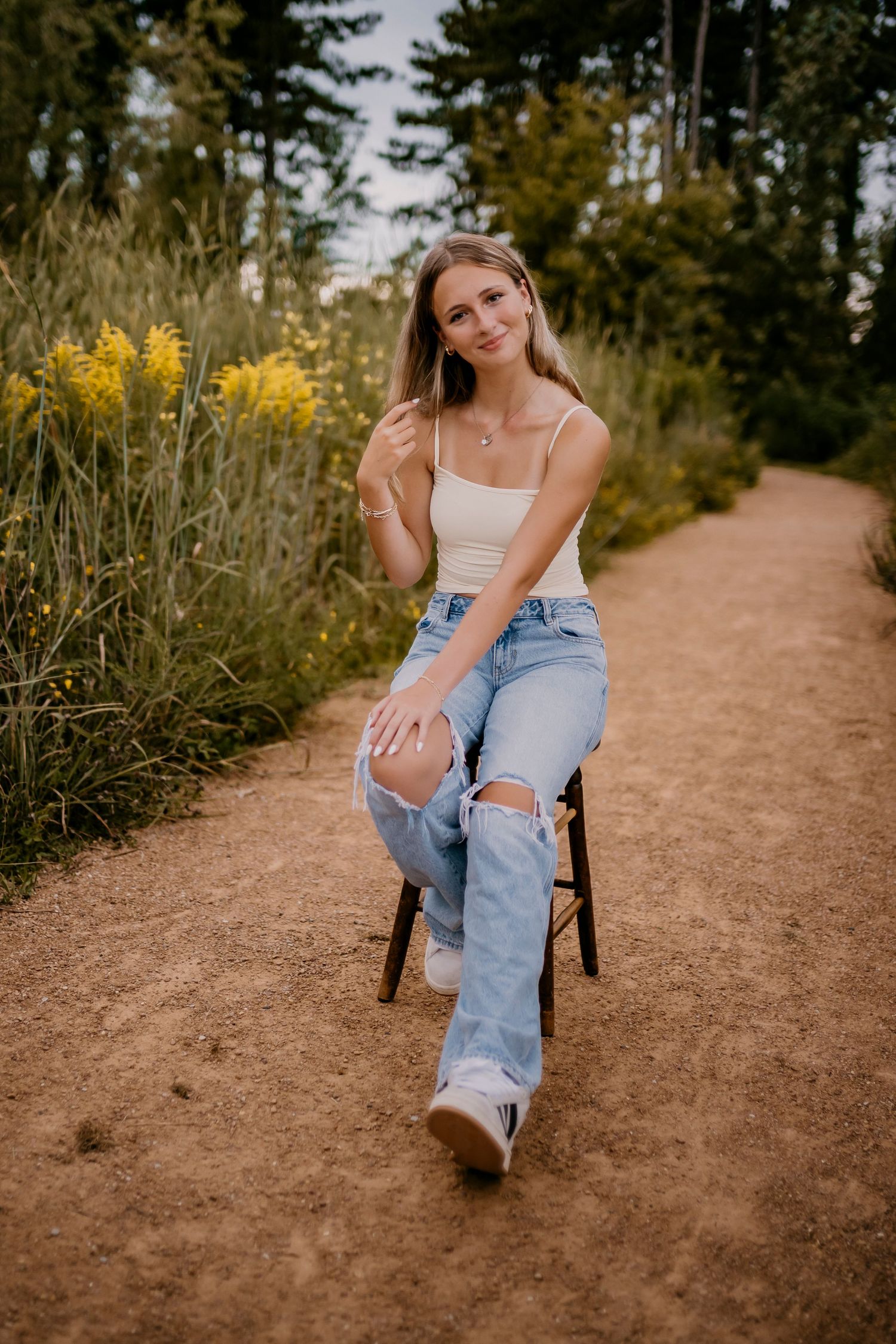 Blonde teen girl in cream tank top and ripped jeans sits on stool along a scenic dirt forest path.