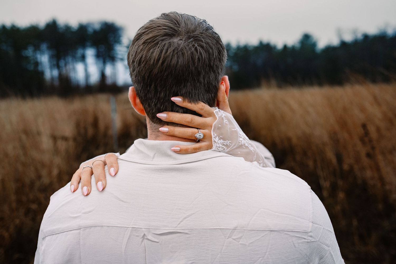 Romantic couple embrace from behind in autumn field, woman wearing diamond engagement ring.