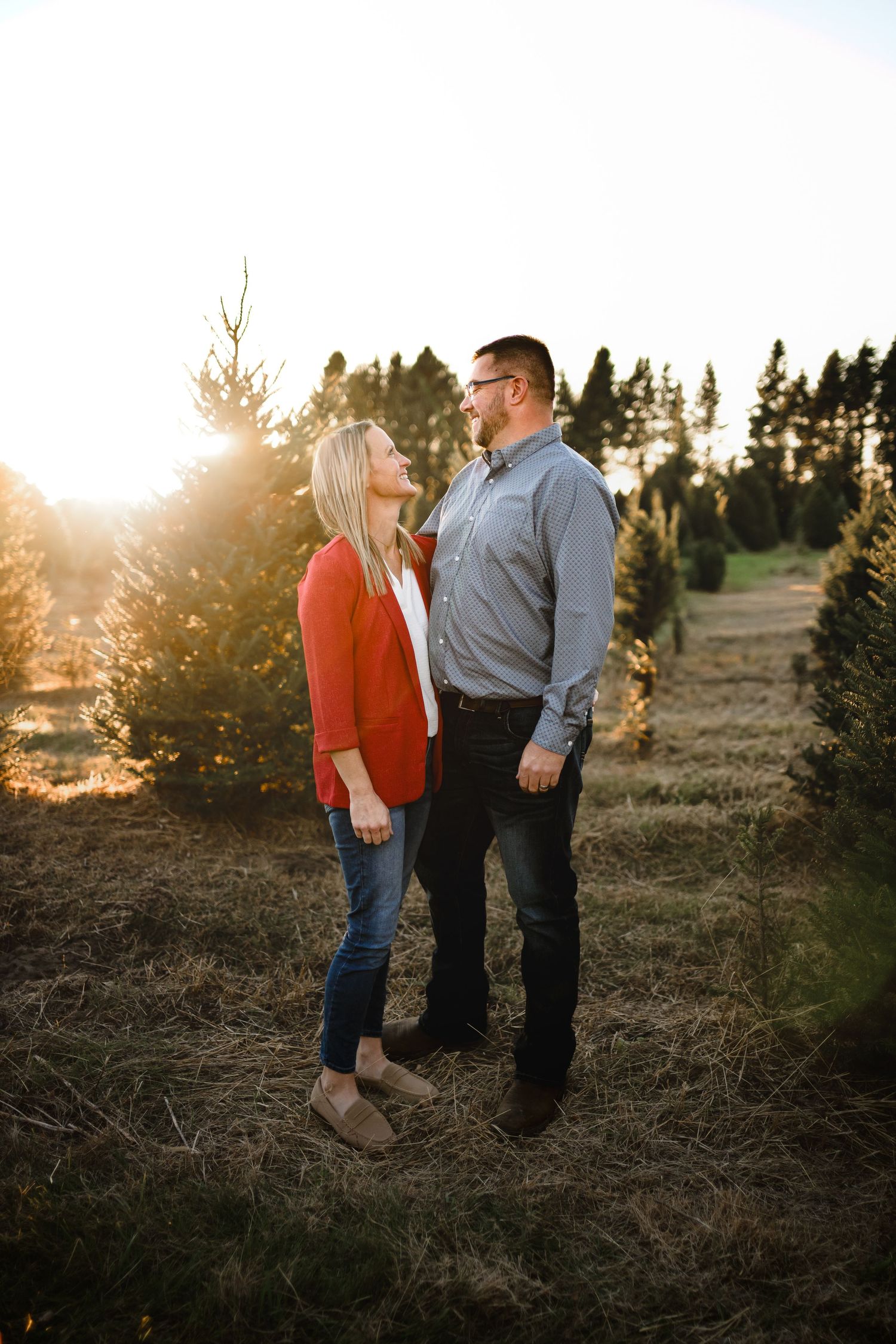 A couple holds hands and gazes at each other at golden hour among Christmas trees on a farm.