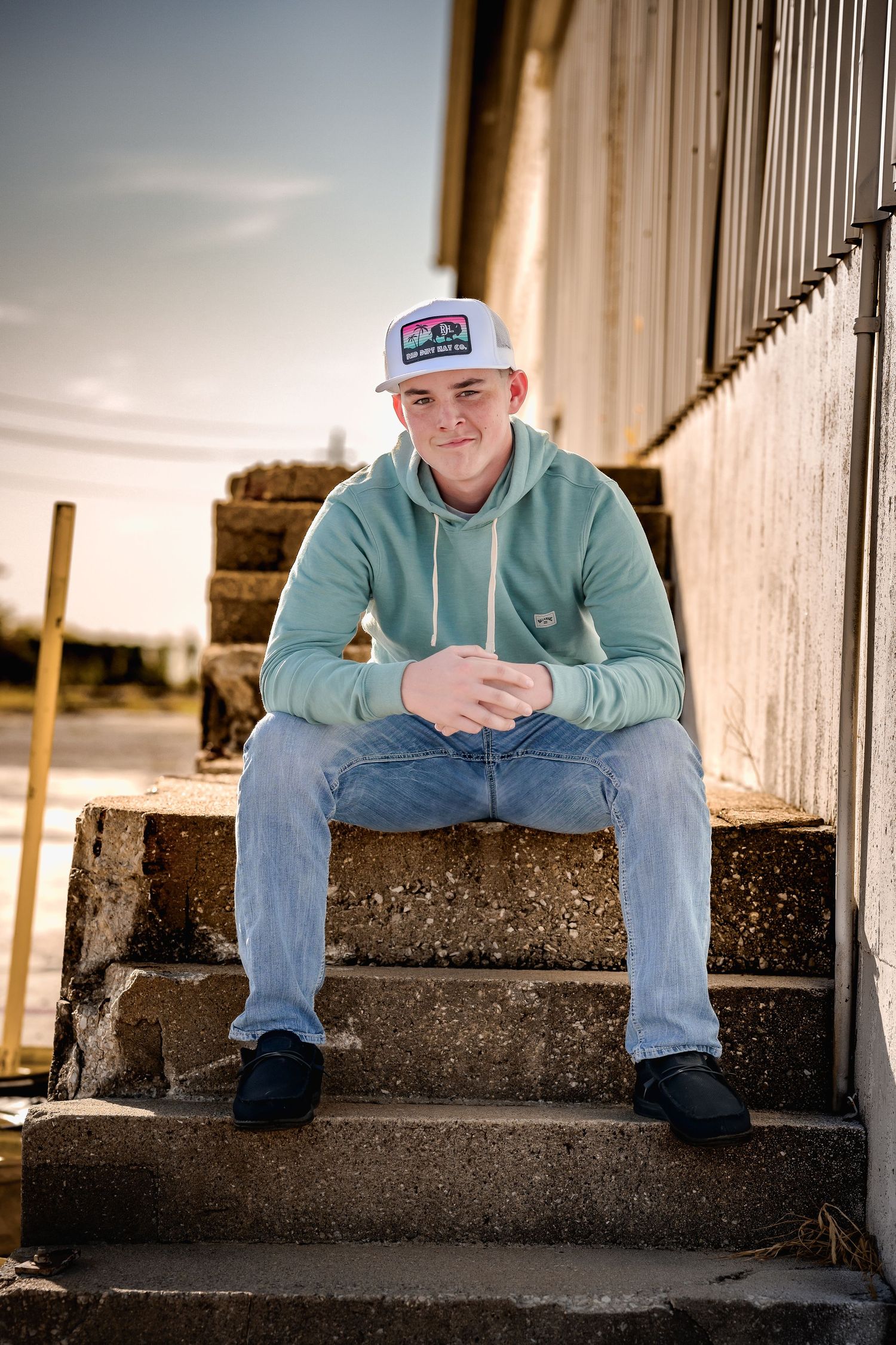 Young man in mint hoodie and trucker hat sitting on concrete steps outdoors.