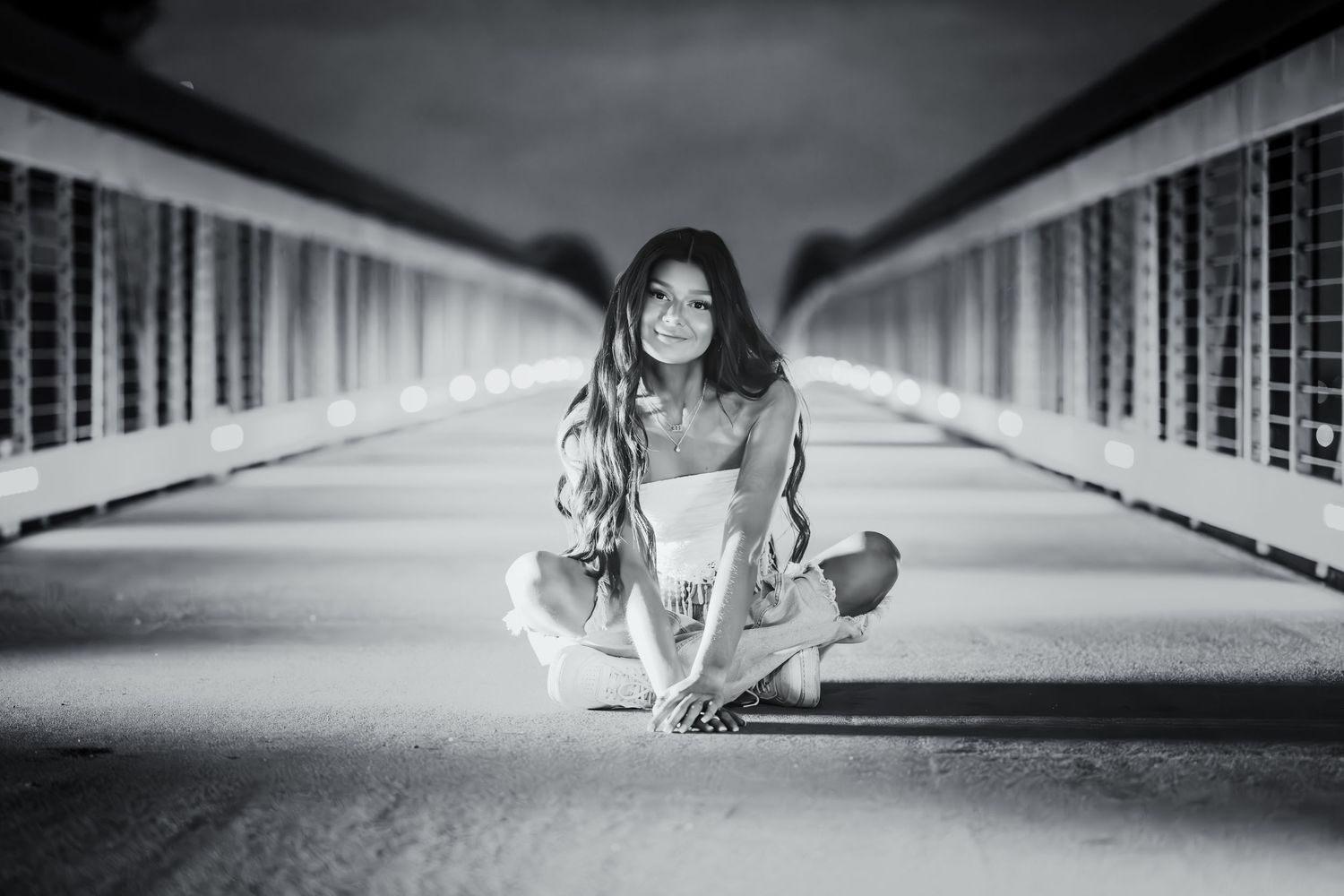 Black and white portrait of young woman sitting cross-legged on a bridge walkway.