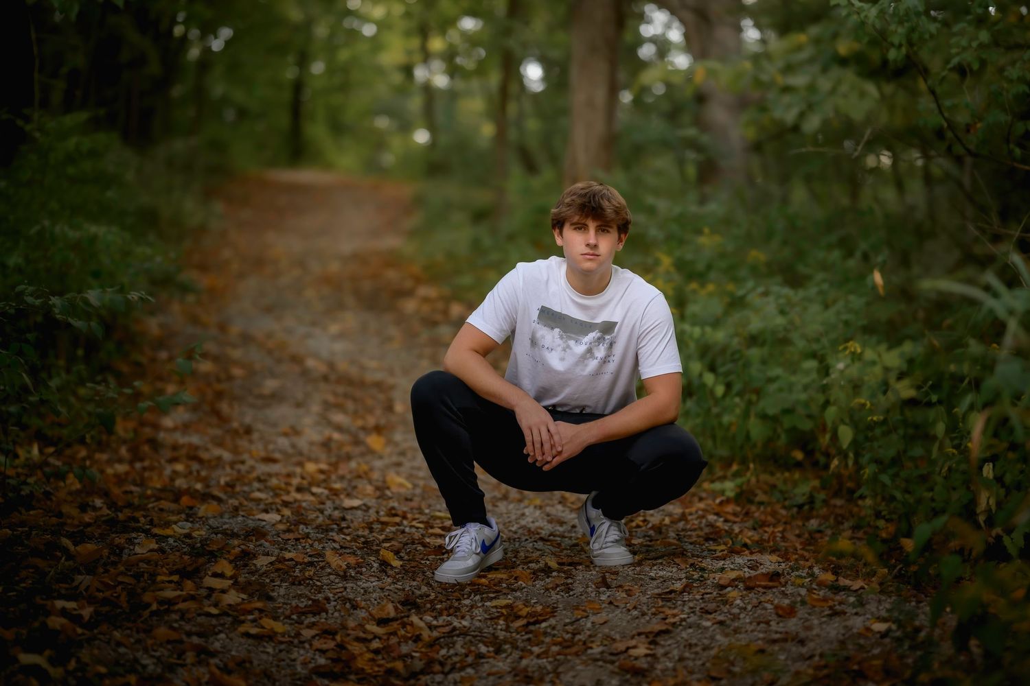 Teen boy in white graphic tee crouching on leaf-covered path in green forest.