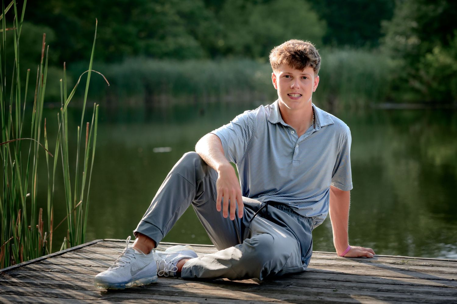 Smiling teenage boy in blue polo sitting relaxed on wooden dock beside calm lake.