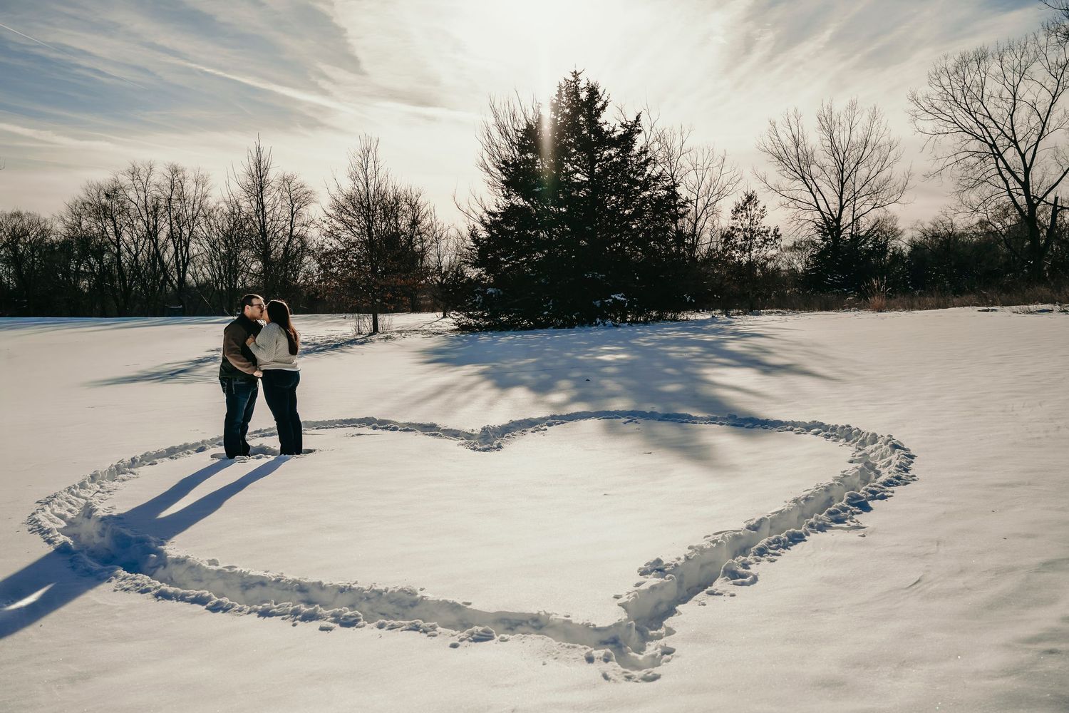 Couple standing inside large heart drawn in snow in sunny winter park landscape.