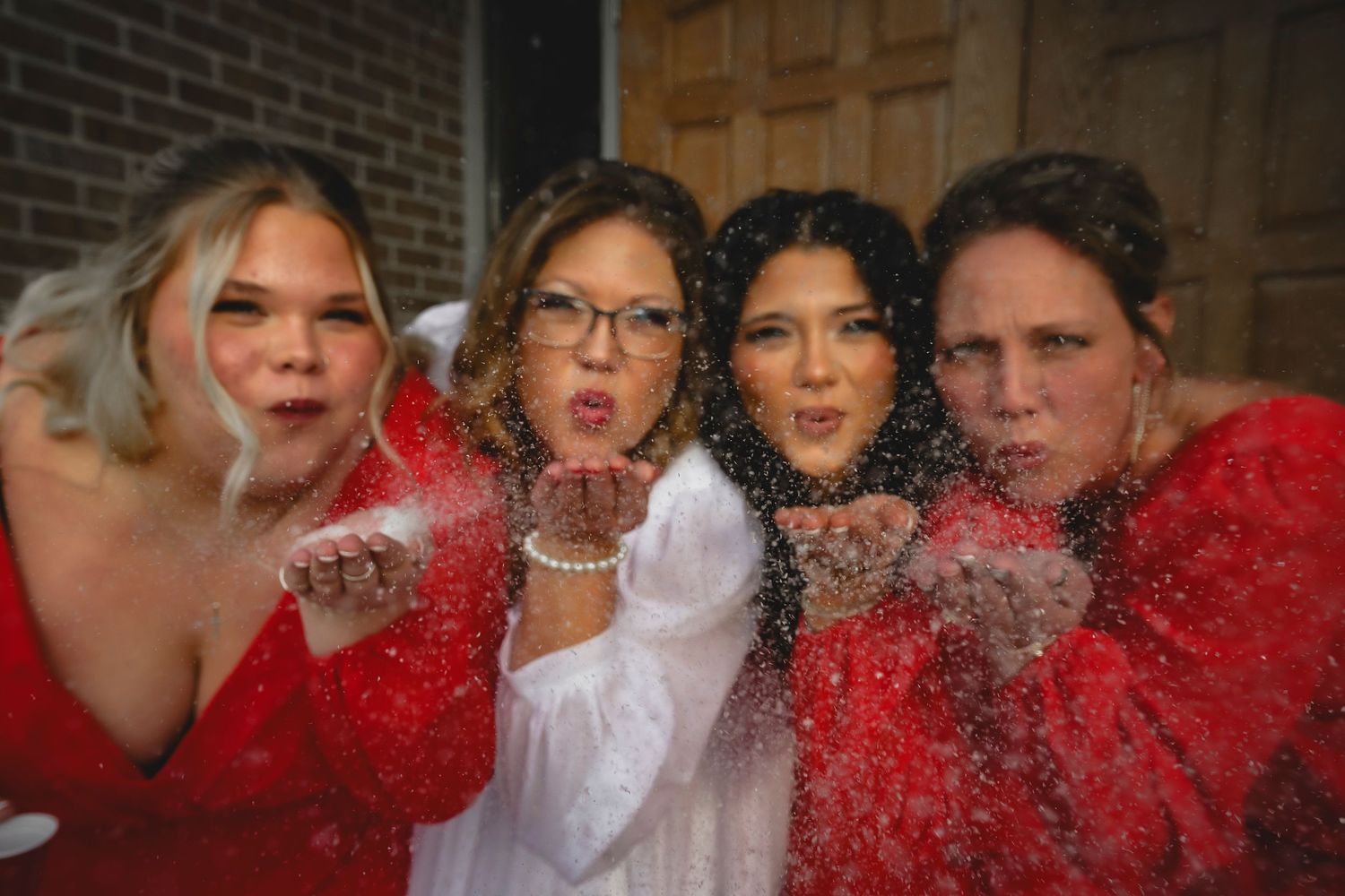 Four women in red and white dresses blowing glitter toward the camera in a festive celebratory photo.