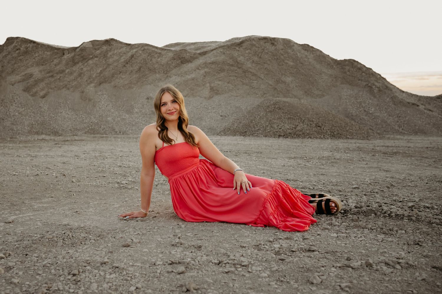 Woman in red maxi dress poses gracefully on rocky desert ground with gravel mounds at sunset.