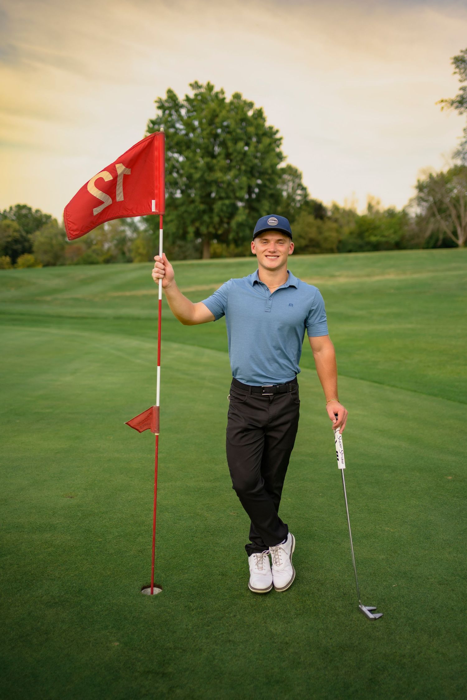 Young male golfer holds red flag and club on lush green golf course at sunset.
