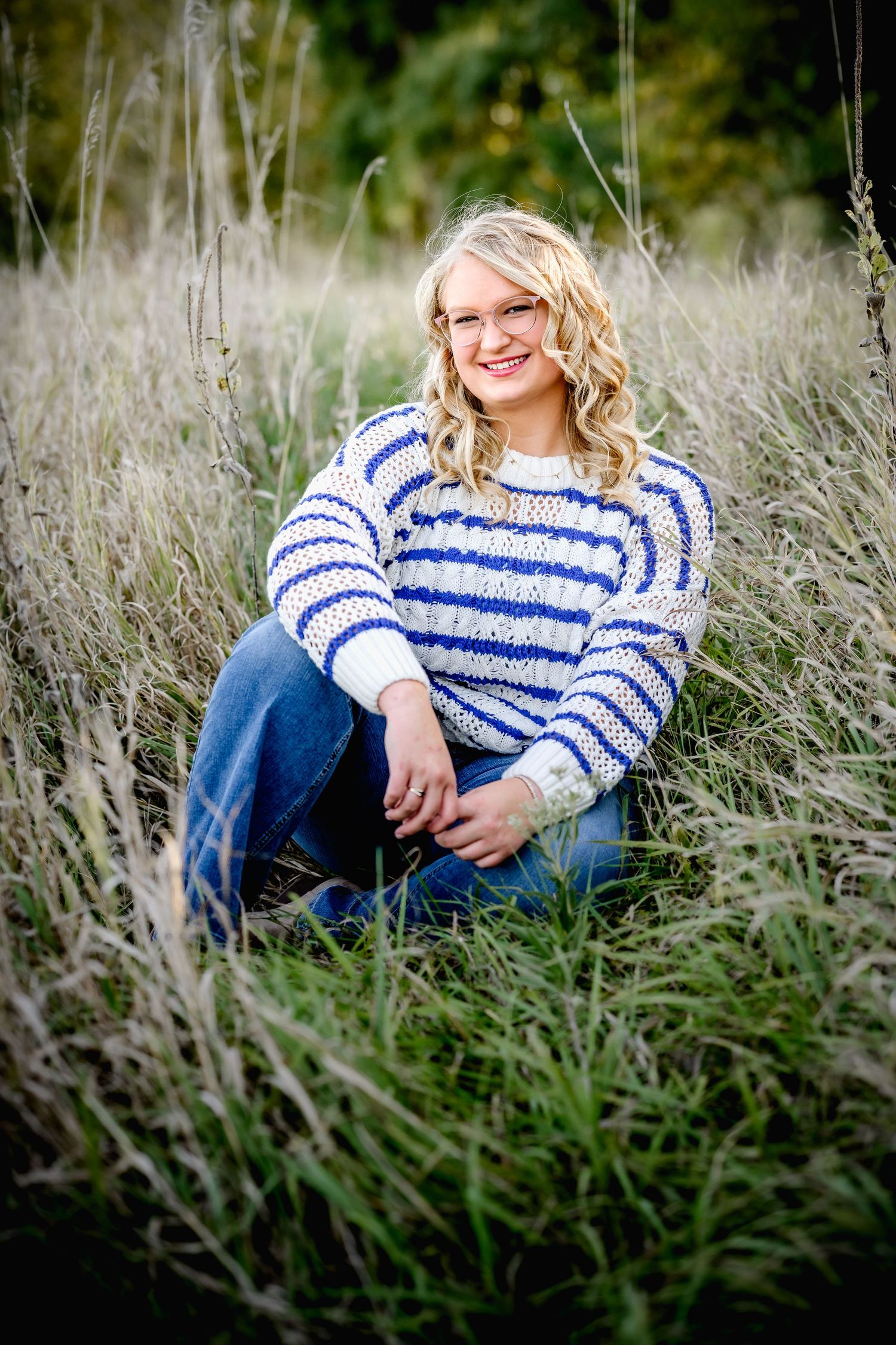 Young blonde woman in blue striped sweater and jeans sitting in tall grass field, smiling outdoors.