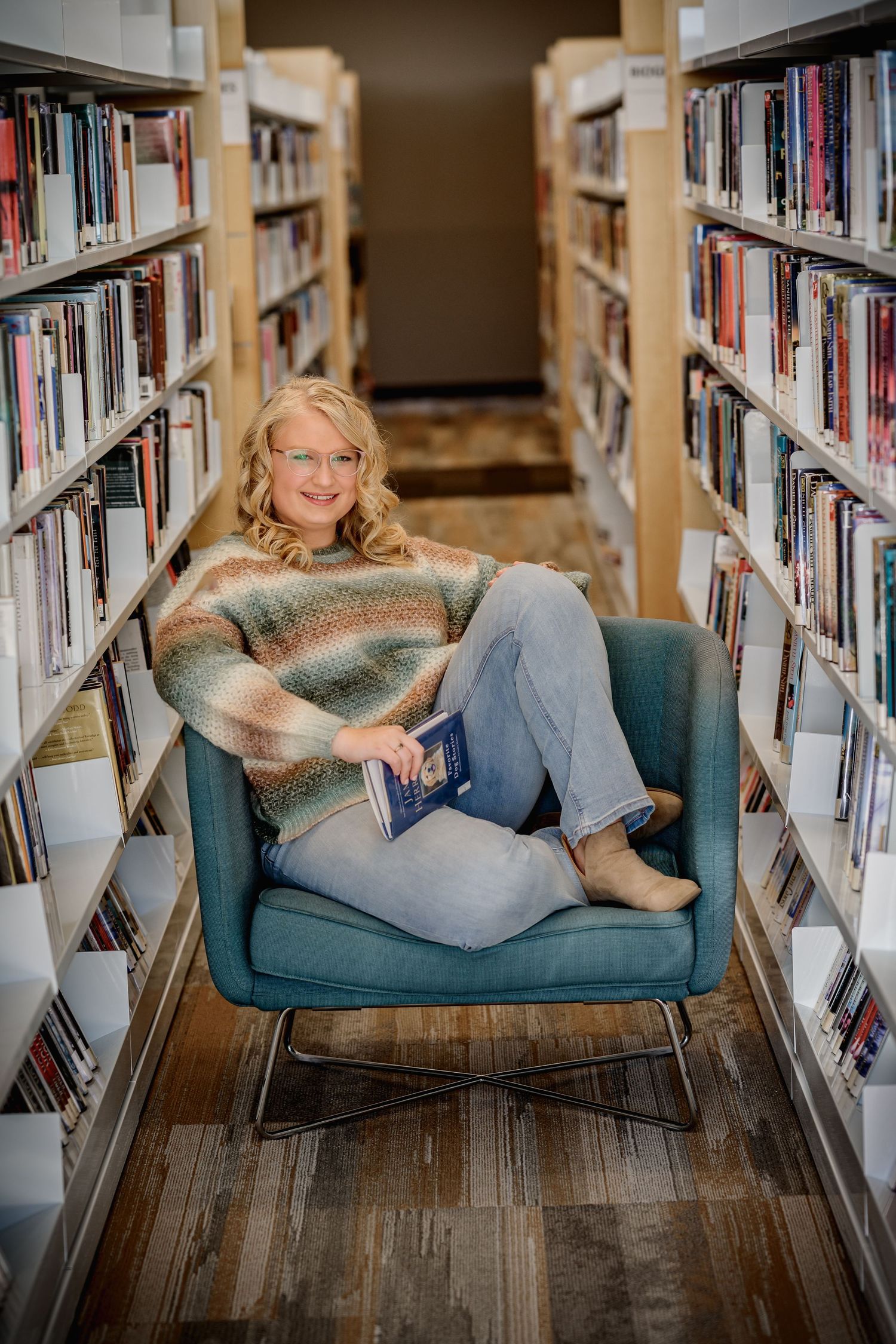 Smiling blonde woman in a cozy sweater sits in a teal chair between library bookshelves holding a book.