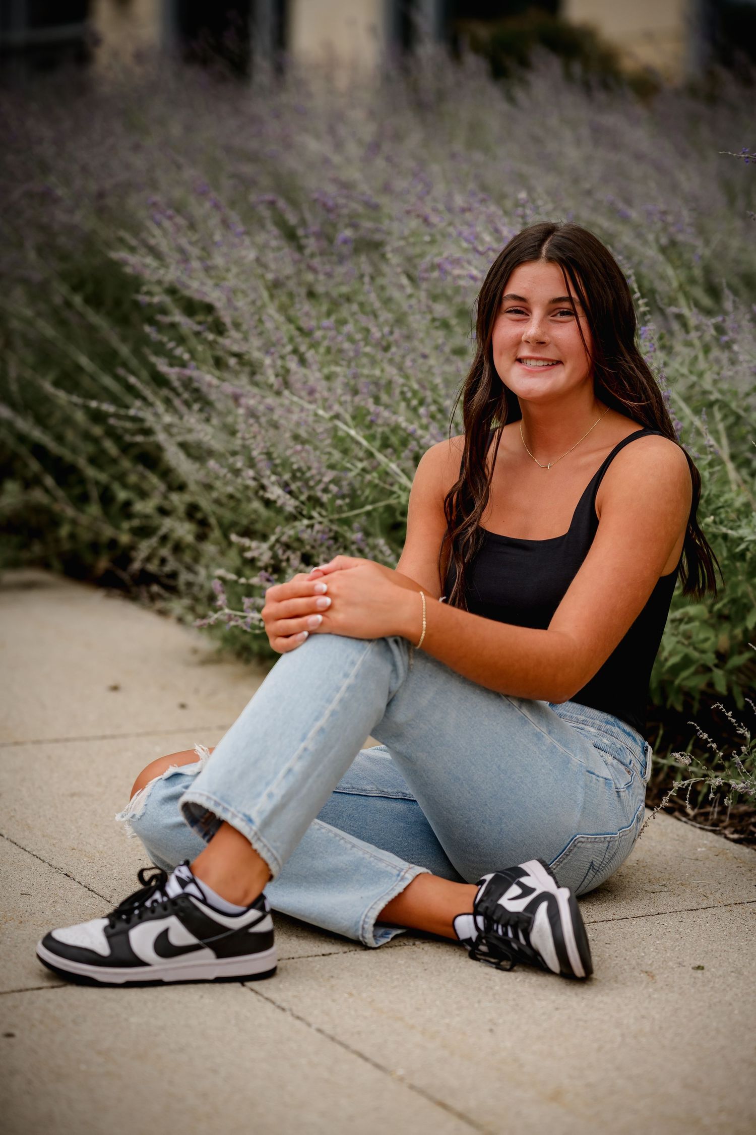 Teen girl in black tank top and light jeans sits on pavement in front of purple lavender bushes wearing Nike Dunks.