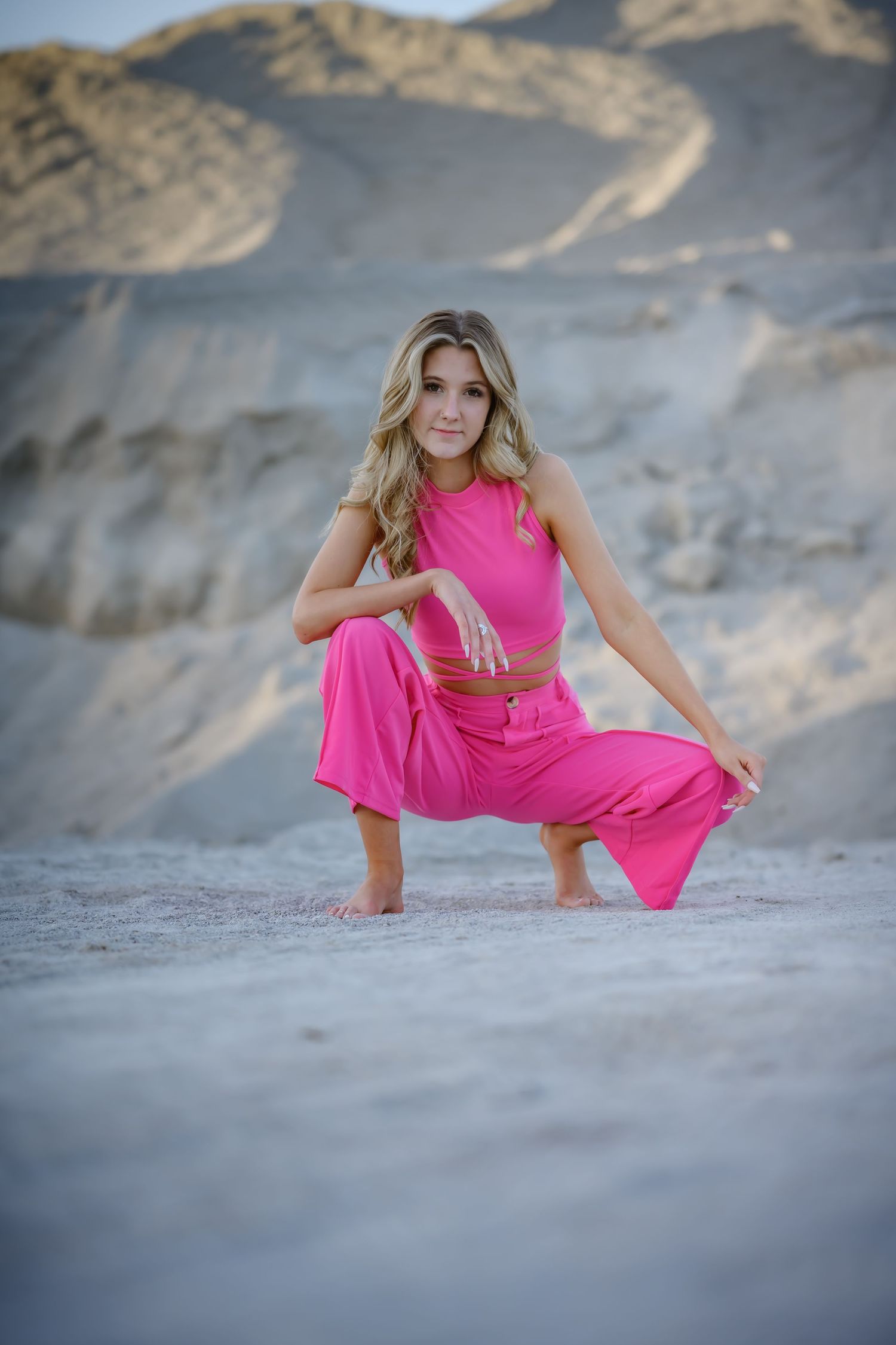 Blonde woman in pink crop top and wide-leg pants crouching in sandy desert landscape.