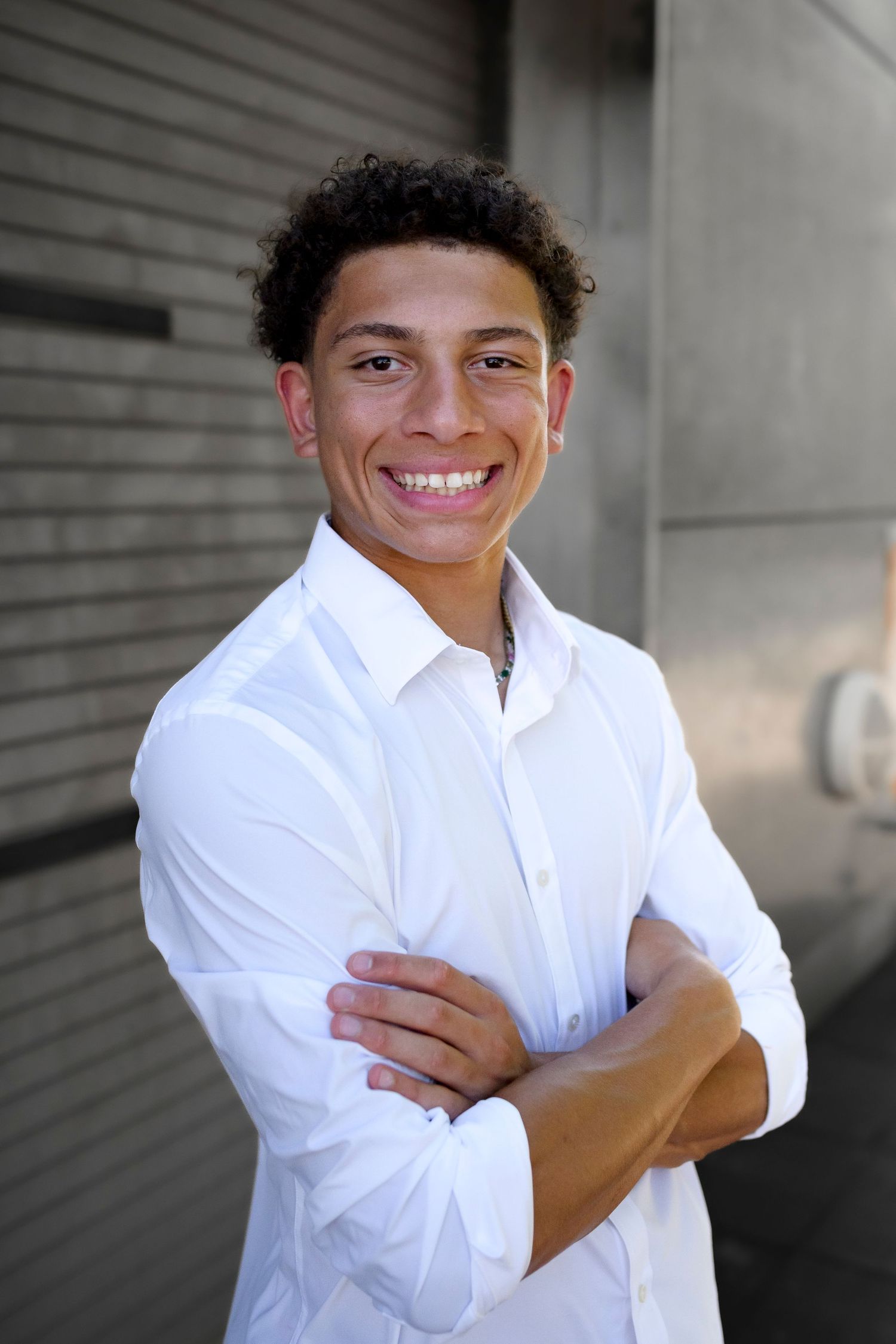 Smiling young man with curly hair stands confidently with arms crossed wearing a white button-up shirt.
