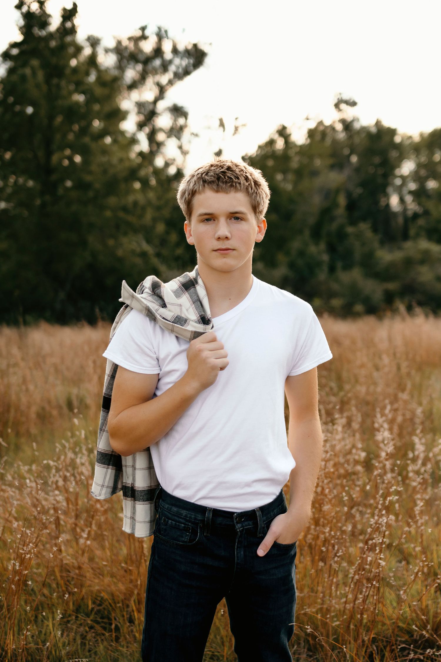 Teen boy in white t-shirt holds plaid flannel jacket over shoulder in golden hour field portrait.