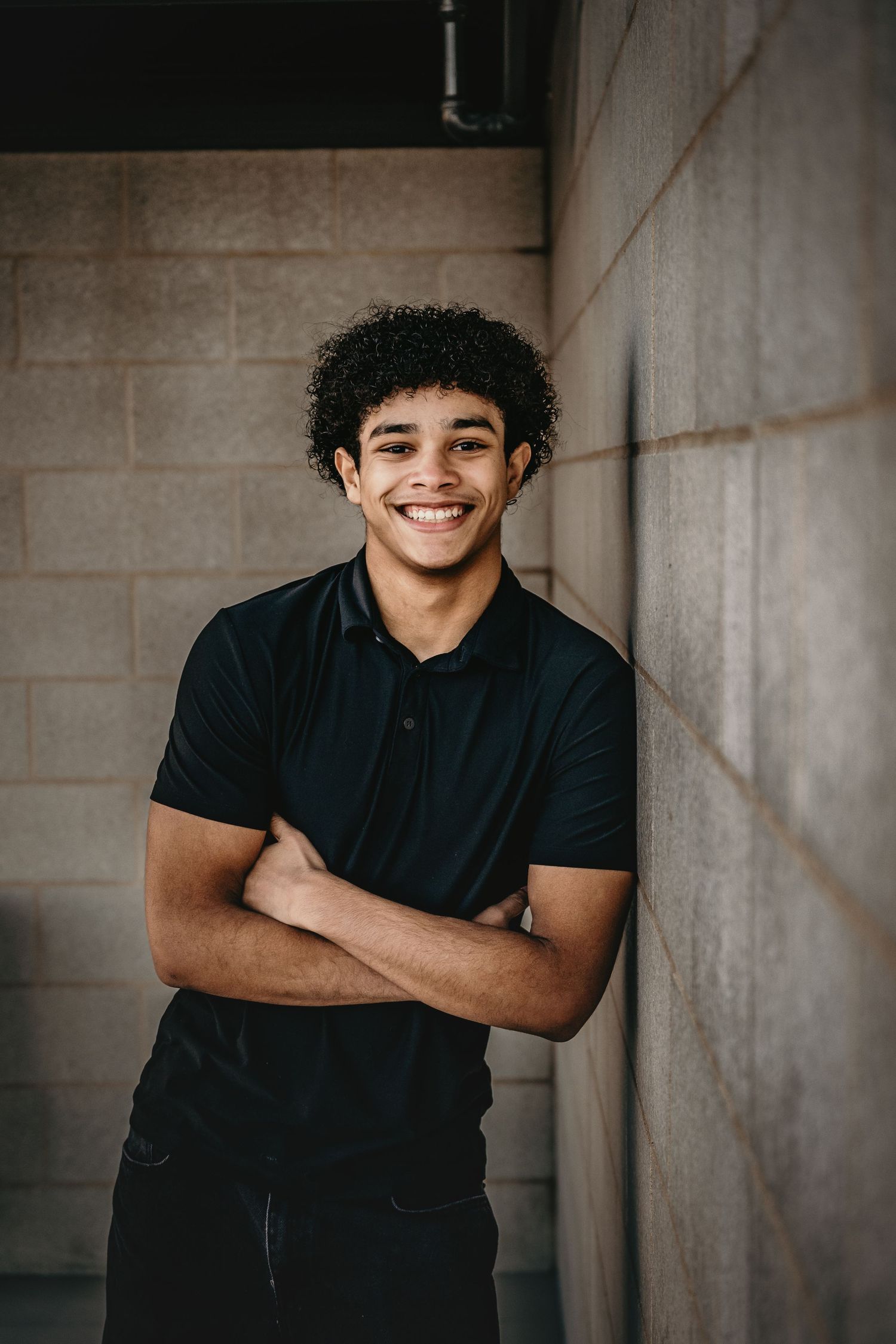 Smiling young man in black polo shirt leans against brick wall with arms crossed.