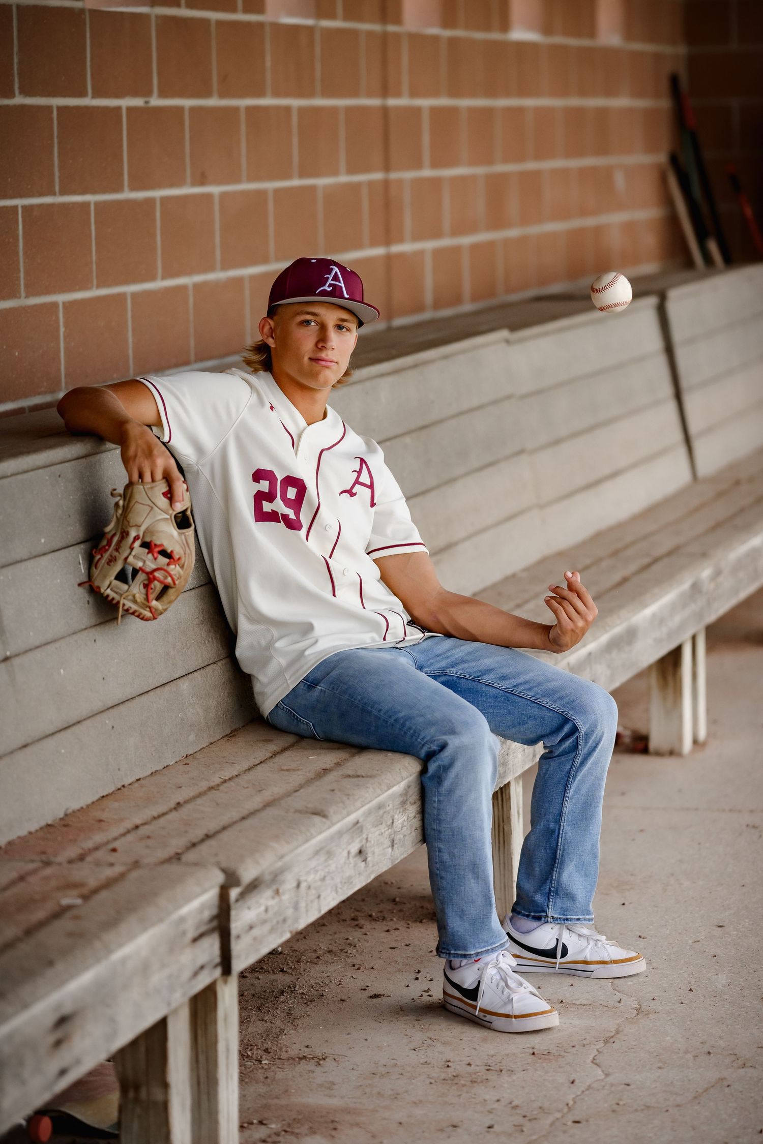 Teen baseball player in uniform sitting in dugout bench tossing a ball with glove.