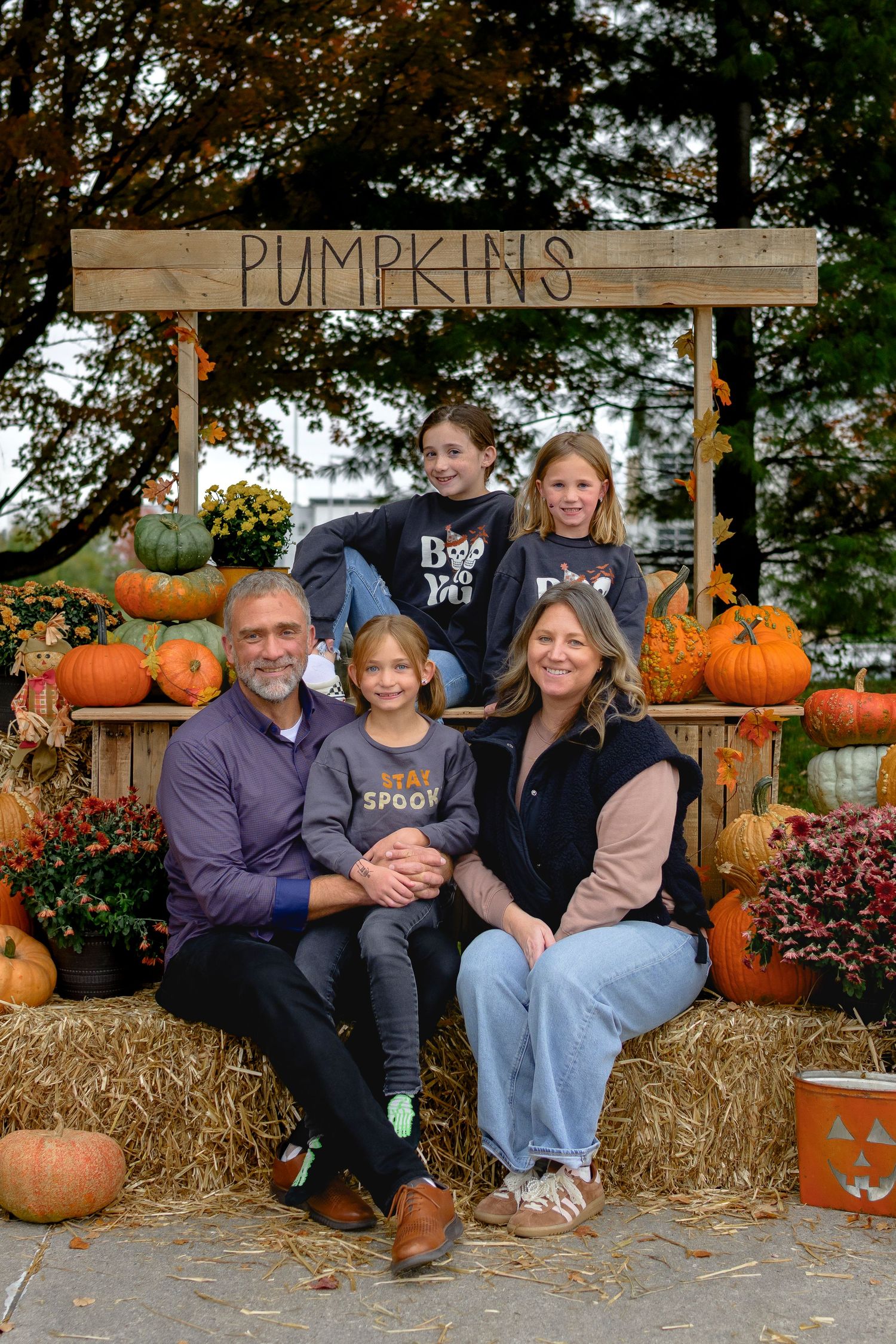 Family of five posing at a fall pumpkin stand surrounded by colorful autumn pumpkins and hay bales.