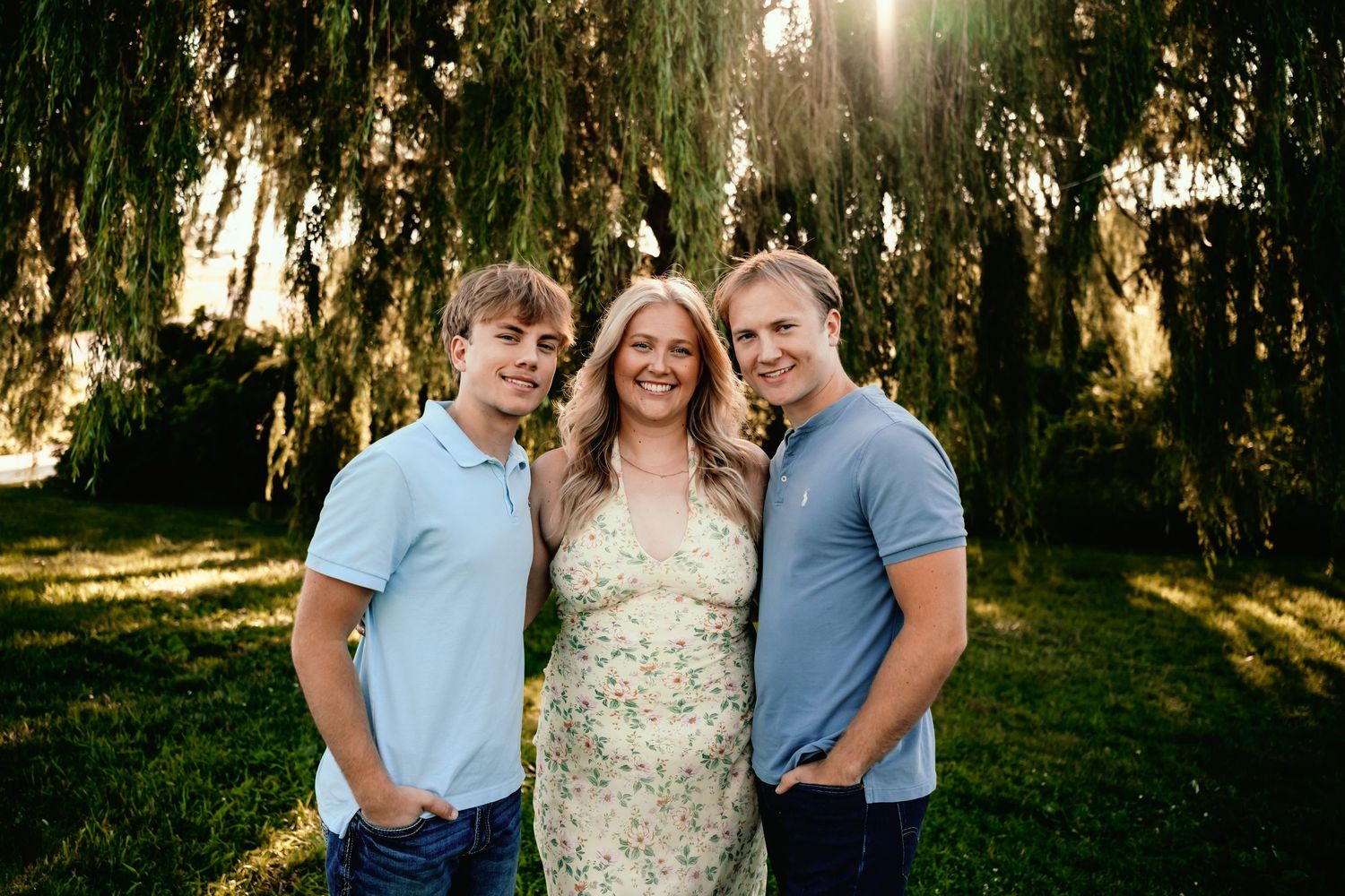 A woman in a floral dress smiles between two young men in blue polos under a willow tree in golden sunlight.