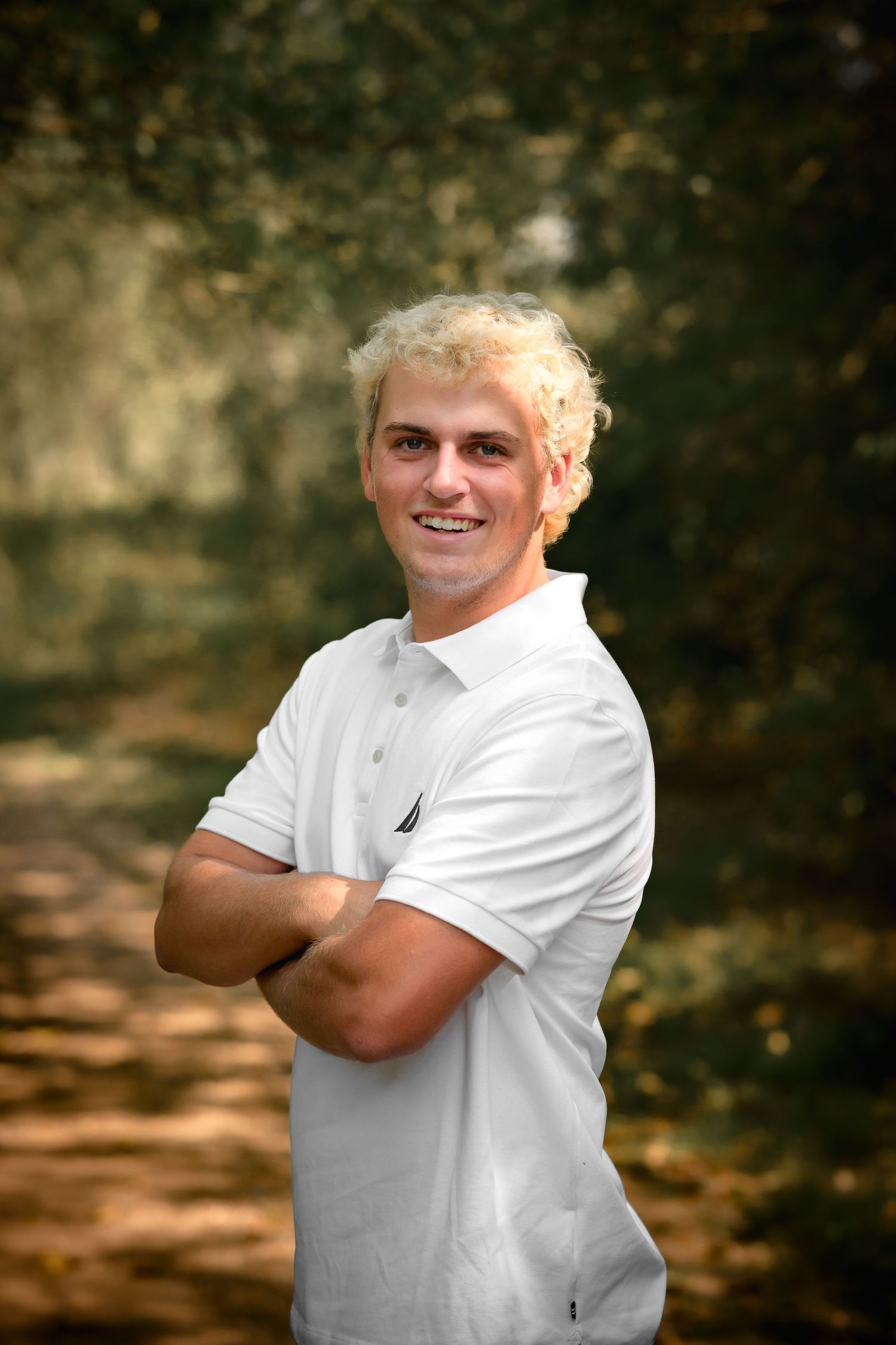 Smiling blond young man in white Nautica polo with arms crossed in wooded setting.