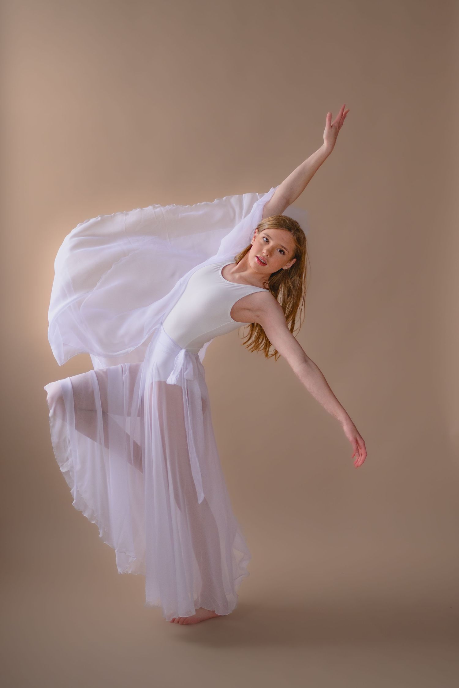 A graceful ballet dancer in a flowing white dress performs an elegant arabesque pose against a beige background.