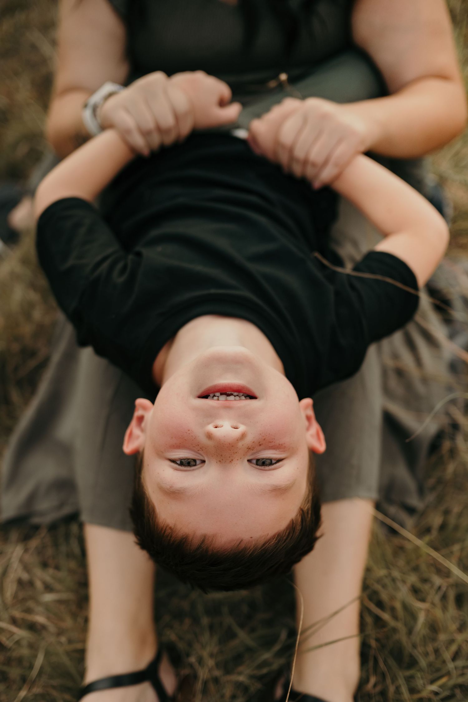 A smiling boy lying upside down on his mother's lap in a grassy field during a family photo session.
