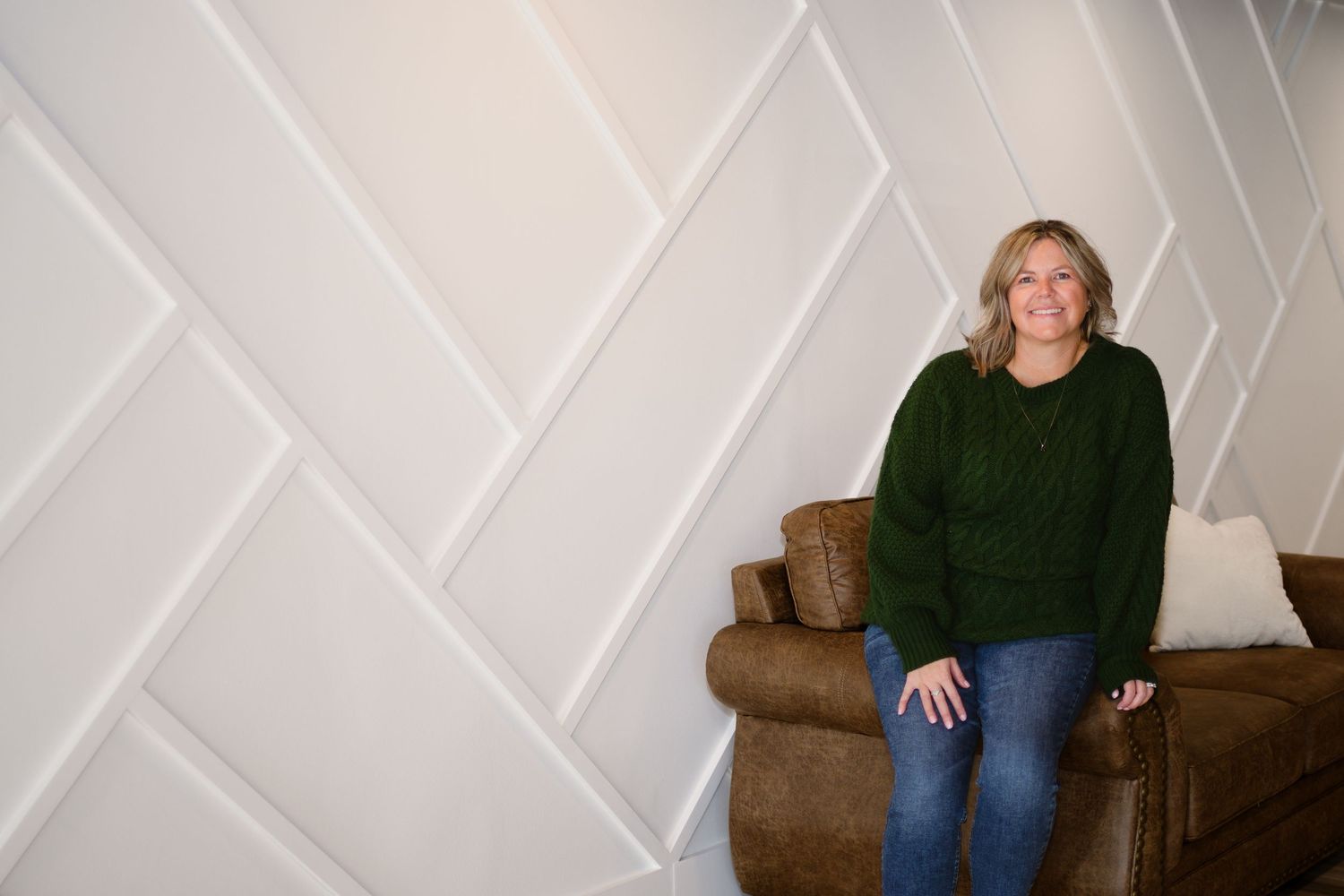 A smiling woman in a green sweater stands beside a couch in front of a geometric white accent wall.