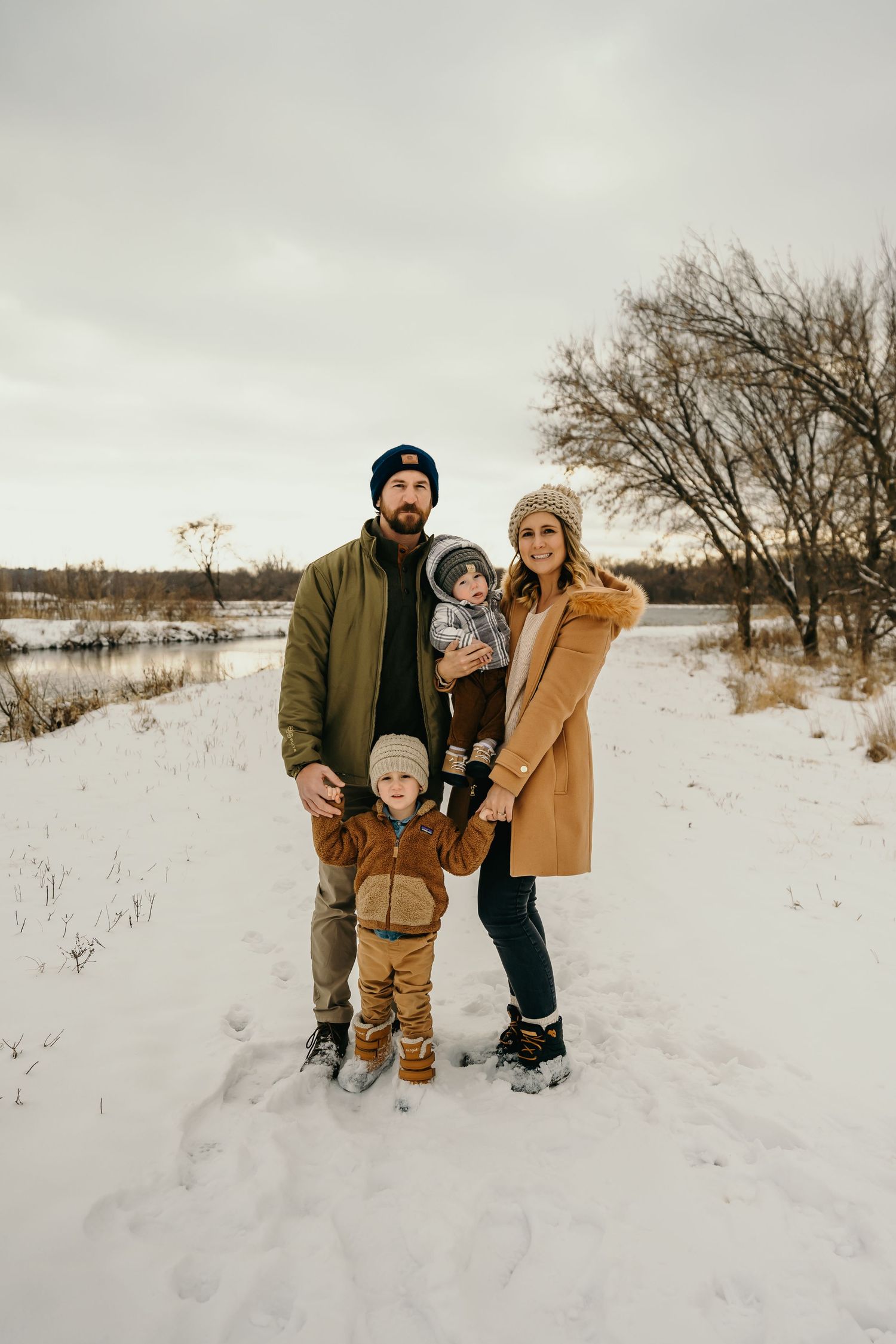 Family of four in winter coats posing in snowy landscape near frozen lake.