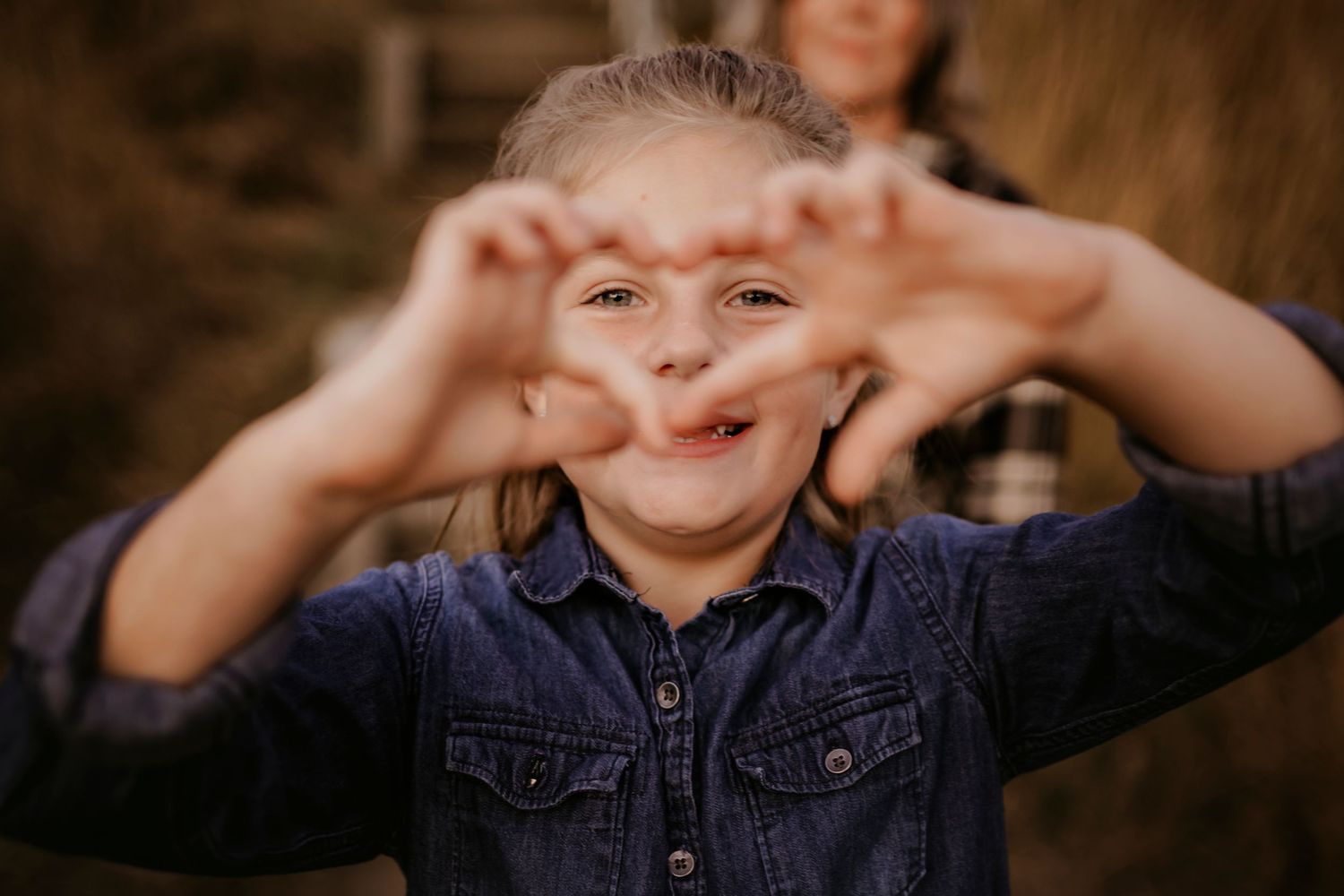 A cheerful young girl in a denim shirt makes a heart shape with her hands toward the camera.
