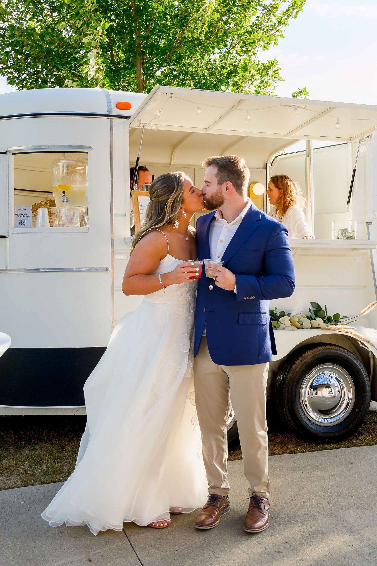 Bride and groom share a kiss beside a white food truck trailer at outdoor wedding reception.