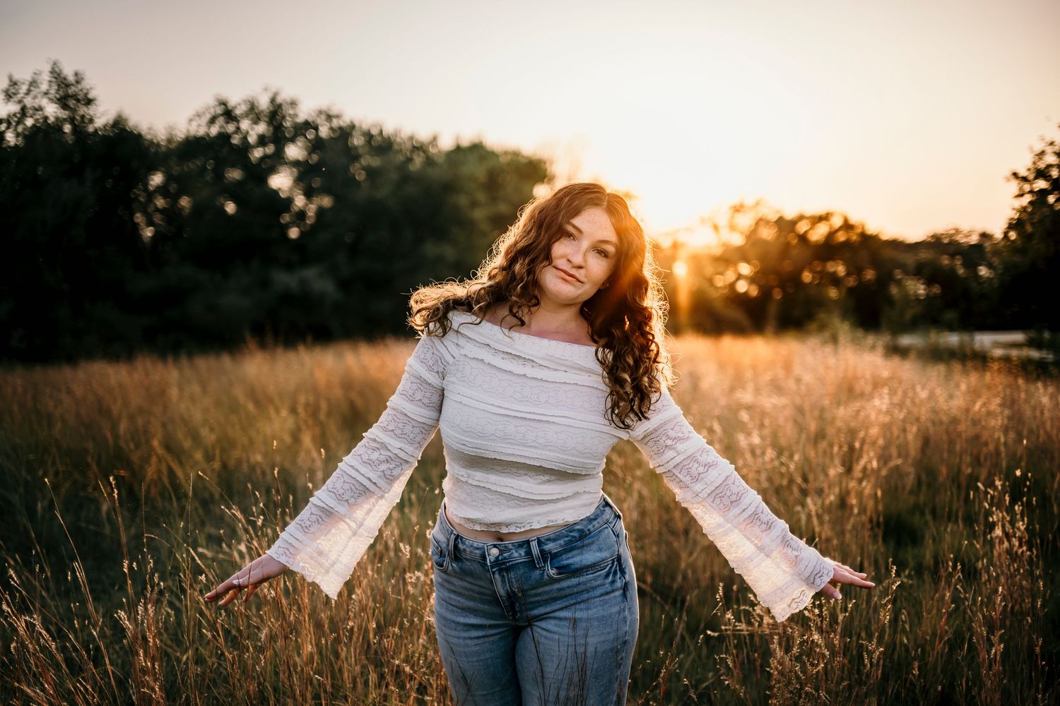Young woman with curly hair poses in golden field at sunset wearing white crop top and jeans.