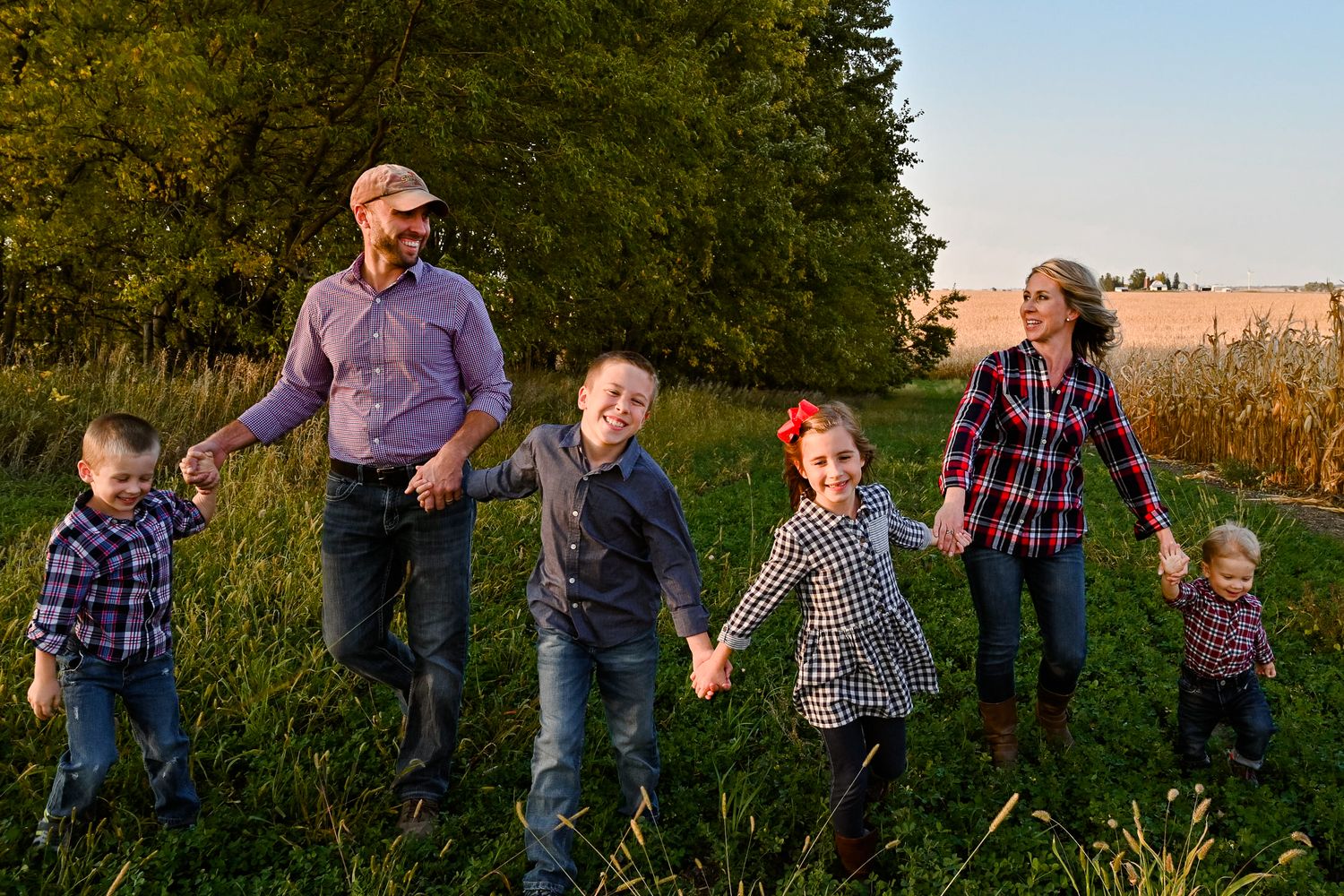 Happy family of six holding hands walking through lush green field at golden hour.