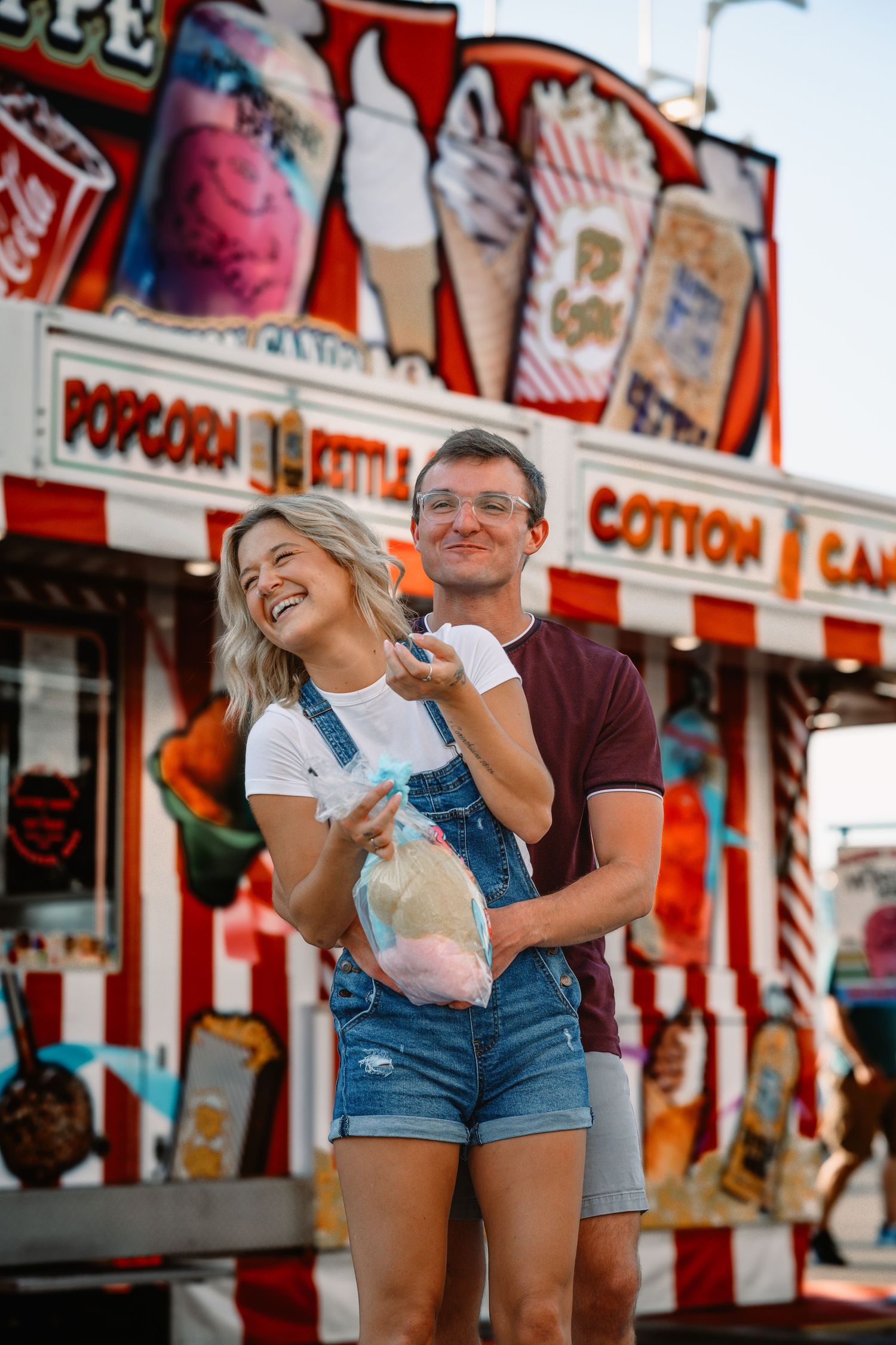 A happy couple laughs and holds cotton candy in front of a colorful carnival food stand at a fair.