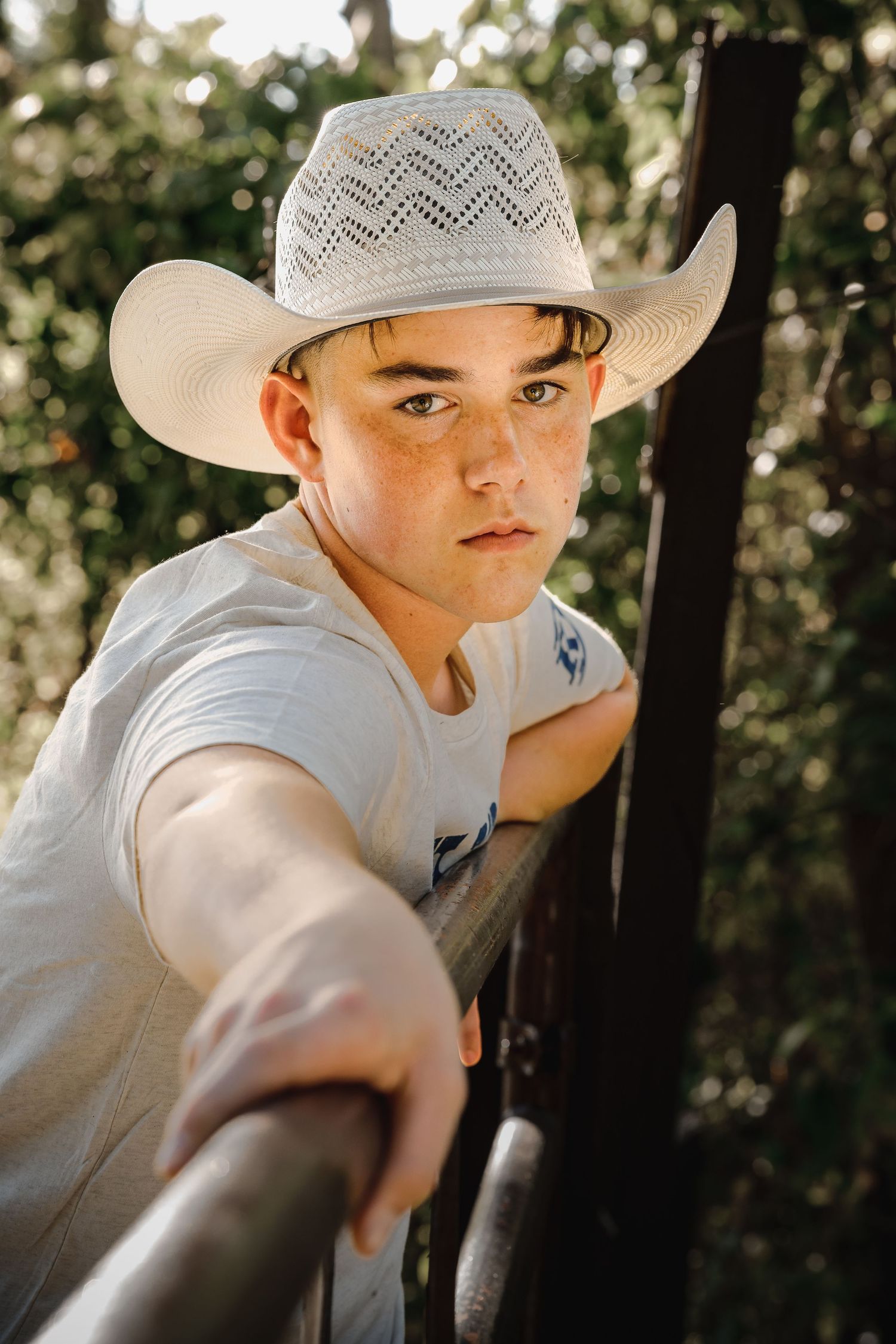 Young teen boy in white cowboy hat leans on a wooden fence rail outdoors in golden afternoon sunlight.