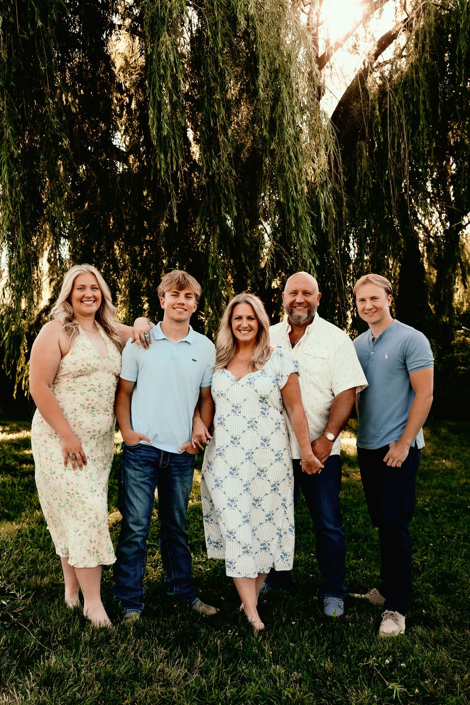 Family of five smiling outdoors under a weeping willow tree during golden hour, dressed in casual summer attire.