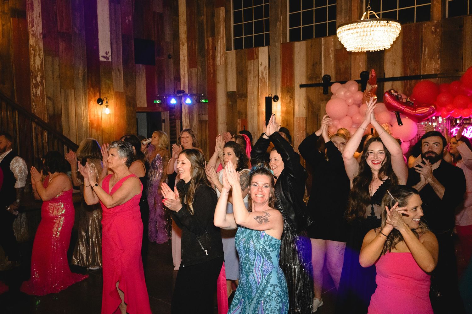 A lively wedding reception crowd claps and dances under warm lights in a rustic barn venue.