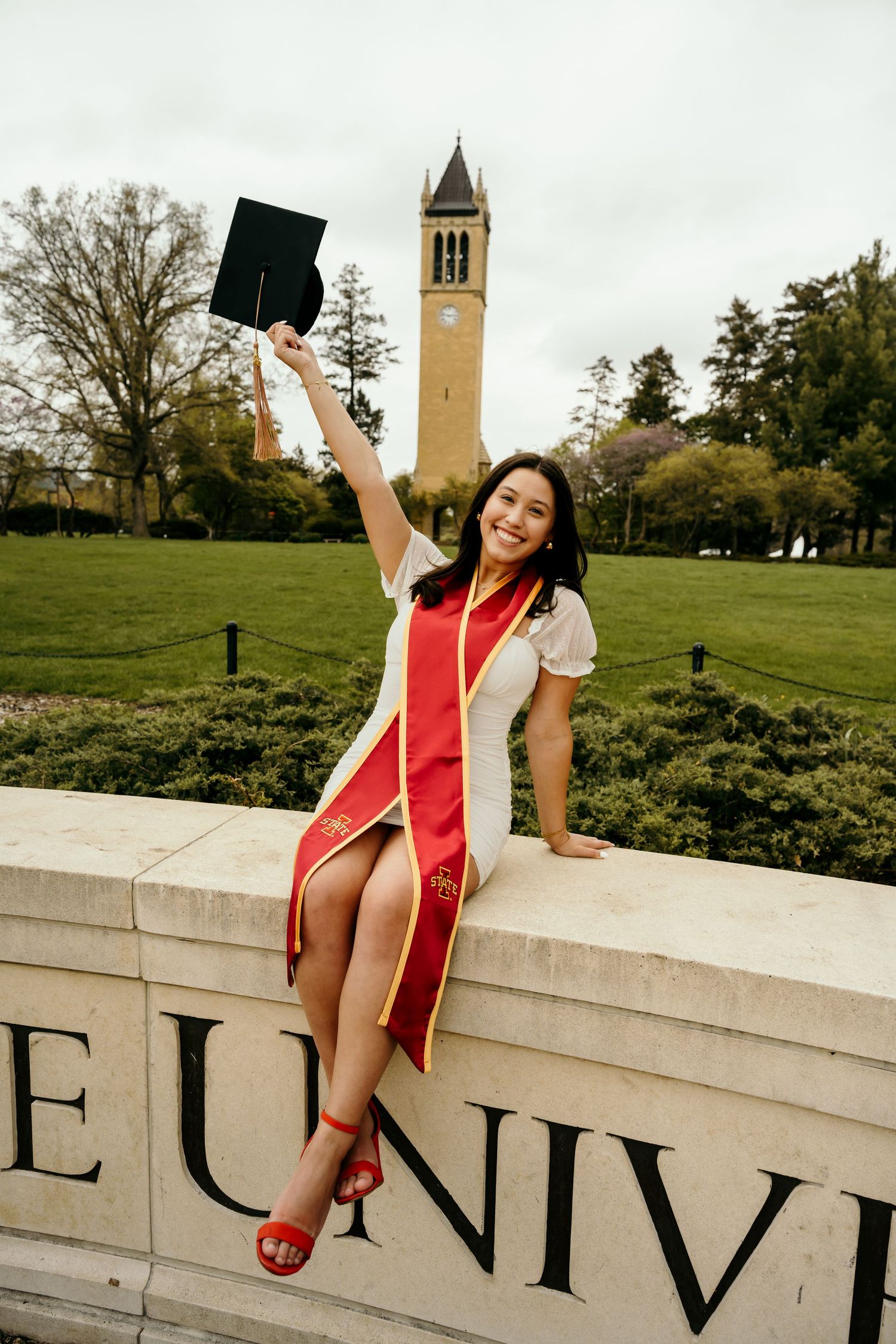 A smiling graduate sits on a university wall holding her cap, wearing a red sash and white dress with a clock tower behind.